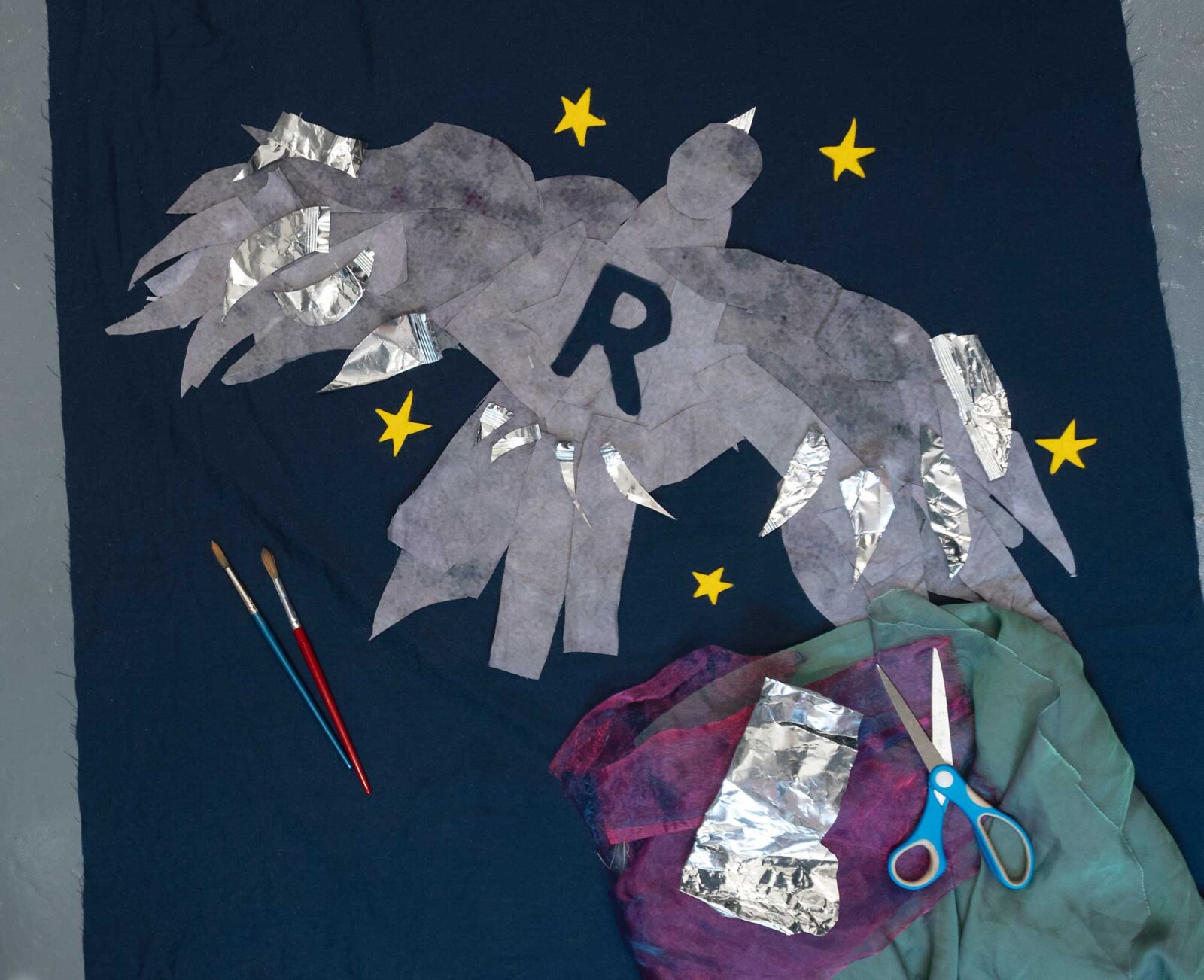 A fabric bird with the letter R on it and yellow stars on it, plus materials such as scissors and fabric to show it was constructed
