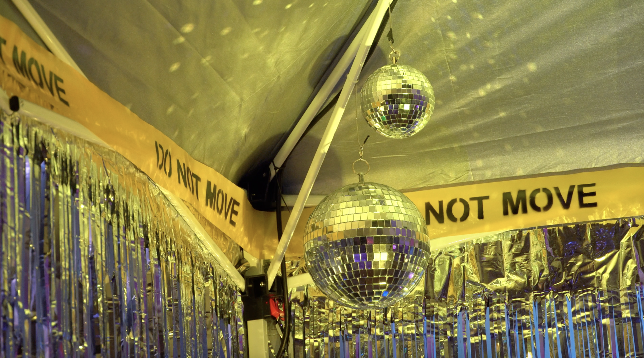 A close up of a top corner of the pop-up mini club. We see DO NOT MOVE hazard tape that lines the top of this tent, with accompanying disco balls that glint and reflect yellow spots onto the top of the fabric