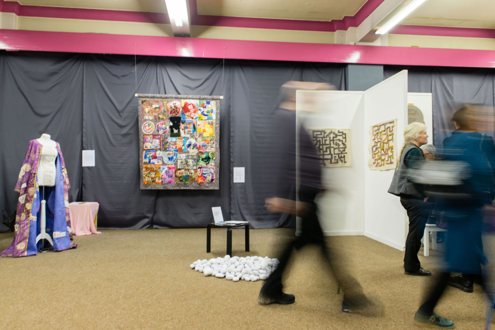 A photo of a blurry person walks through an exhibition space with a mixed material collage suspended on the wall as well as a coat on display and abstract patterned prints