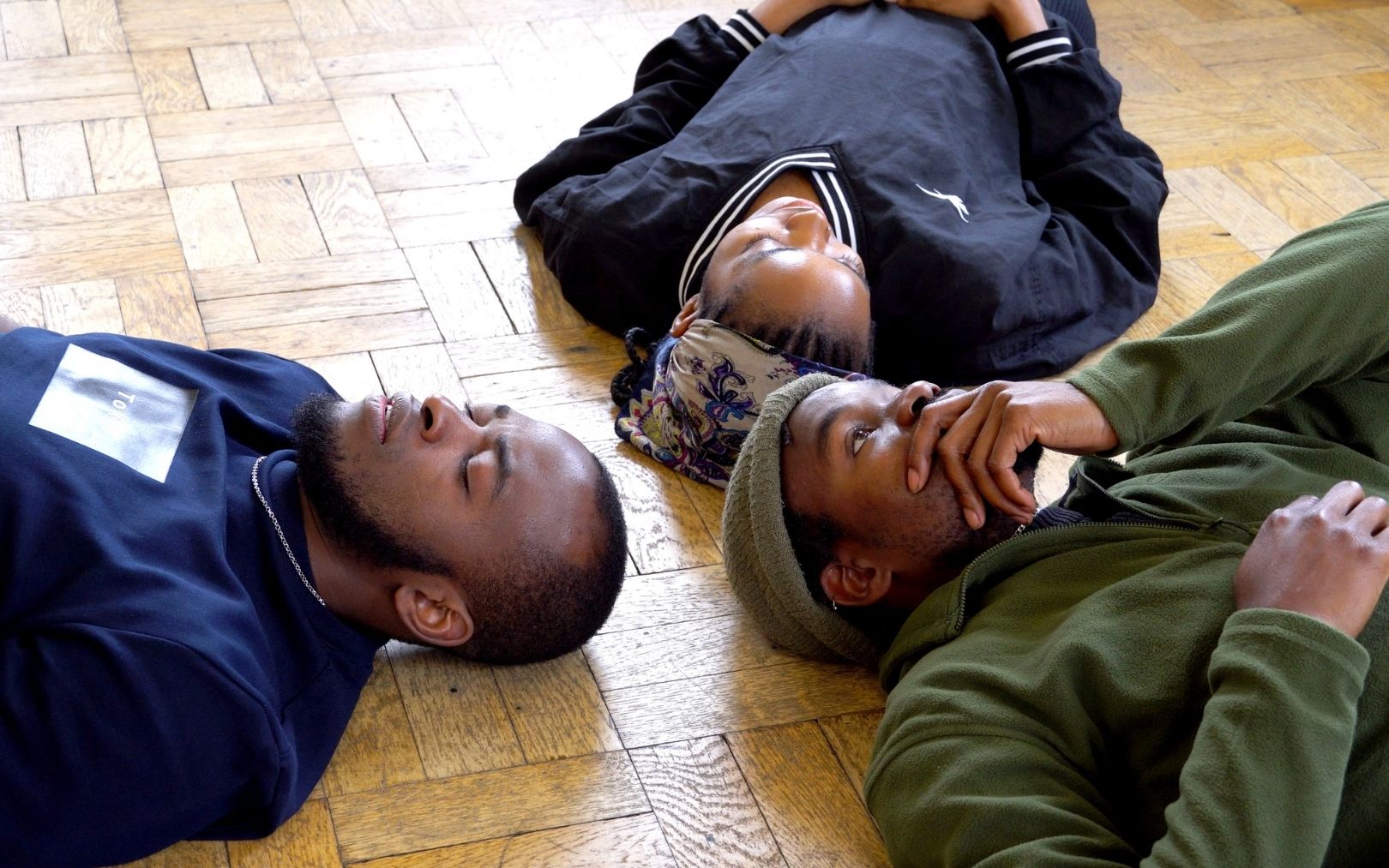 Taiwo is a Black male in his late 20s. He has a short beard and stubble. He wears a green beanie and a green fleece, black jean trousers, and trainers Alya is Black woman in her early 20s. She is wearing a black jersey and black trousers with black converse shoes. She is wearing a blue and white headwrap over braided hair Abdullah is a Black male in his early 30s, he has short black hair and a short shaped beard He is wearing a blue shirt that says Tokyo and a silver chain He is wearing grey checkered trousers