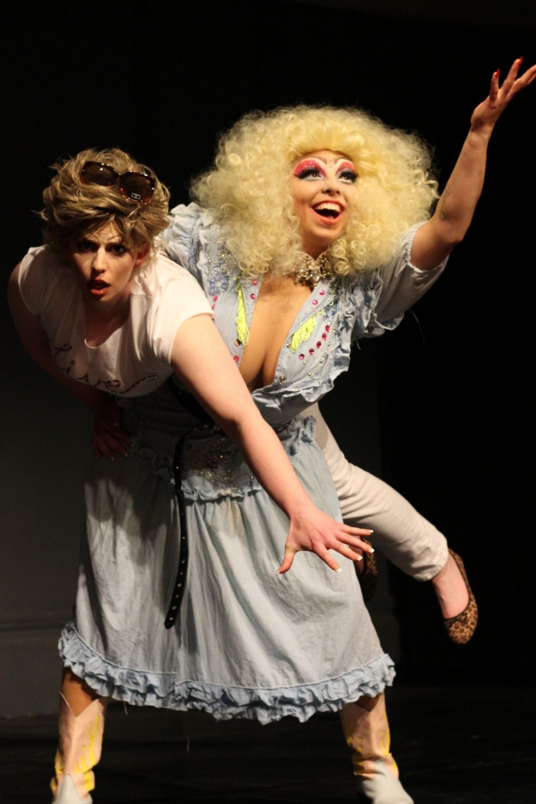 Dolly, wearing a bright blonde curly wig (in the style of Dolly Parton) with red eyeshadow, lifts Karen up under her arm at her waist. Dolly’s arm is raised in celebration. Karen looks concerned and lowers her arm to stabilise herself.