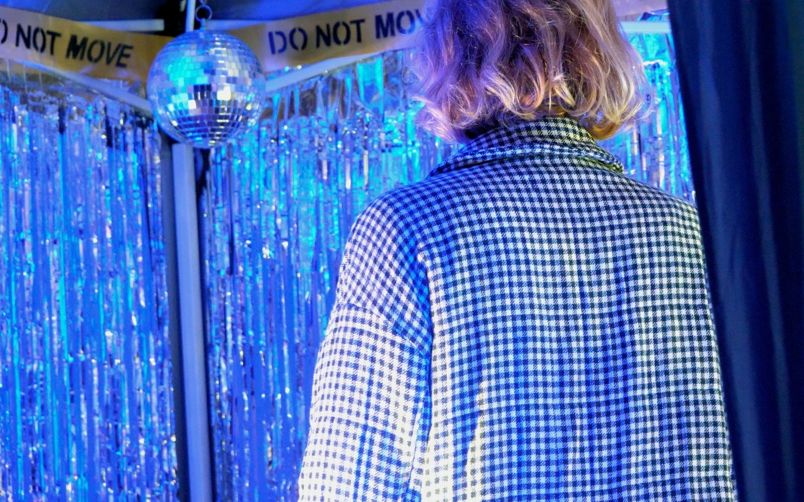A close crop photograph of the back of a person’s torso and head, who faces the inside of the pop-up mini-club. The person has shoulder length blonde hair with a black and white chequed blazer. They are looking into the tent, with foil strip curtains illuminated in blue, a disco ball and the same DO NOT MOVE hazard tape.
