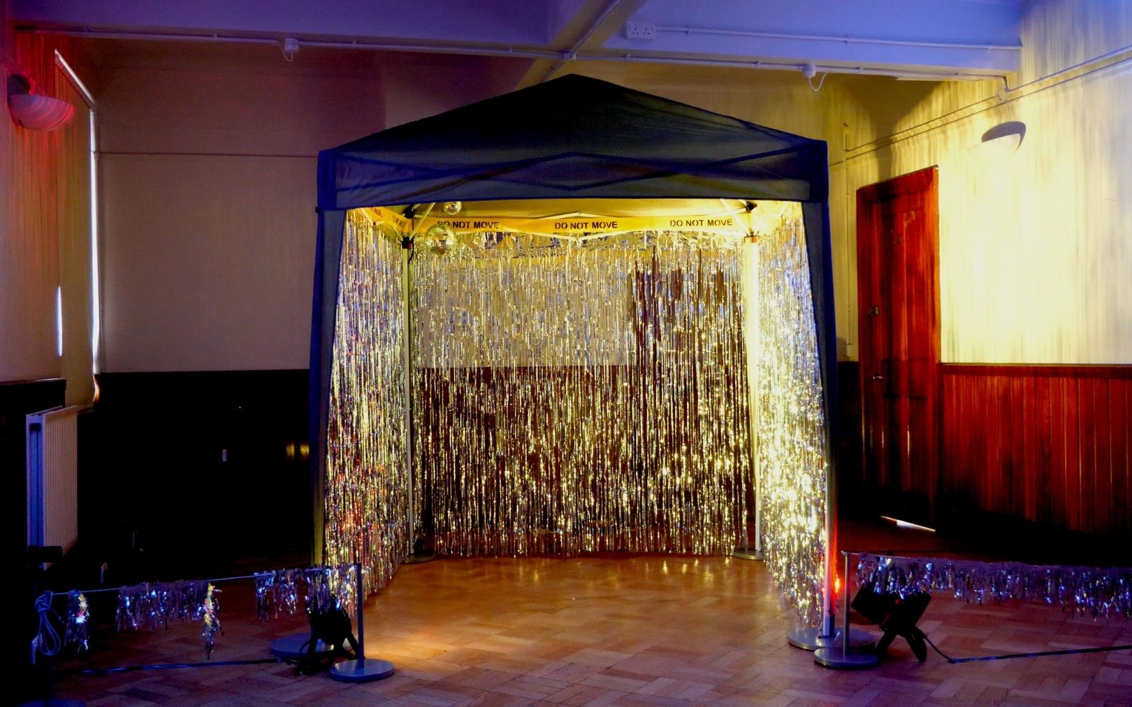 This photograph shows a dimly lit room with a pop-up mini club in the centre of the frame. Foil strip curtains fringe the back and side walls of the structure, illuminated in yellow. DO NOT MOVE hazard tape lines the top of this tent, with accompanying disco balls hanging up inside. Fringed barriers flank the tent structure