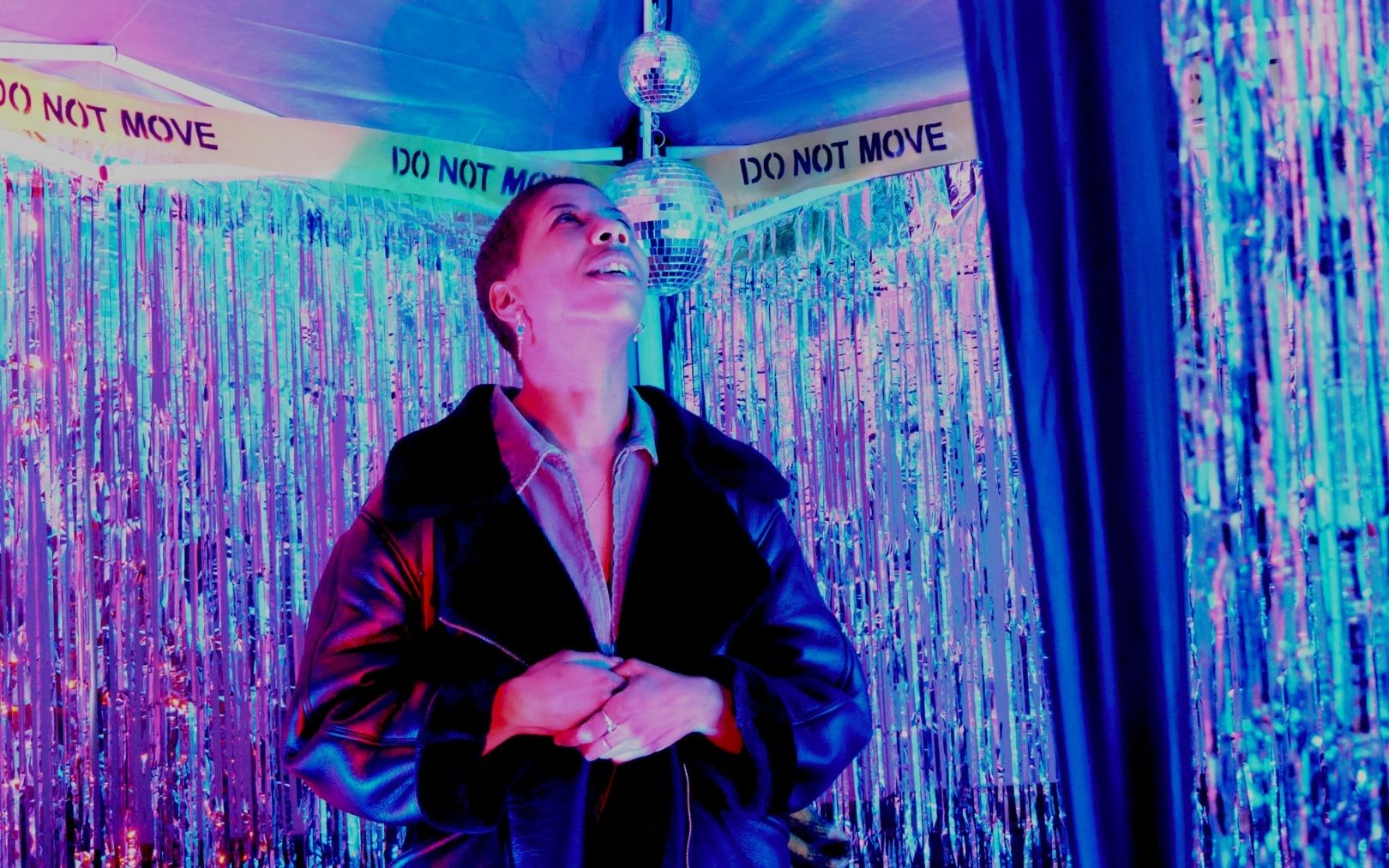 A photograph of a person who appears to be looking up in wonder, inside the disco. They have a black leather jacket and short cropped hair, illuminated in pink. The foil fringe strip curtains look shimmering in blues, pinks and purples.
