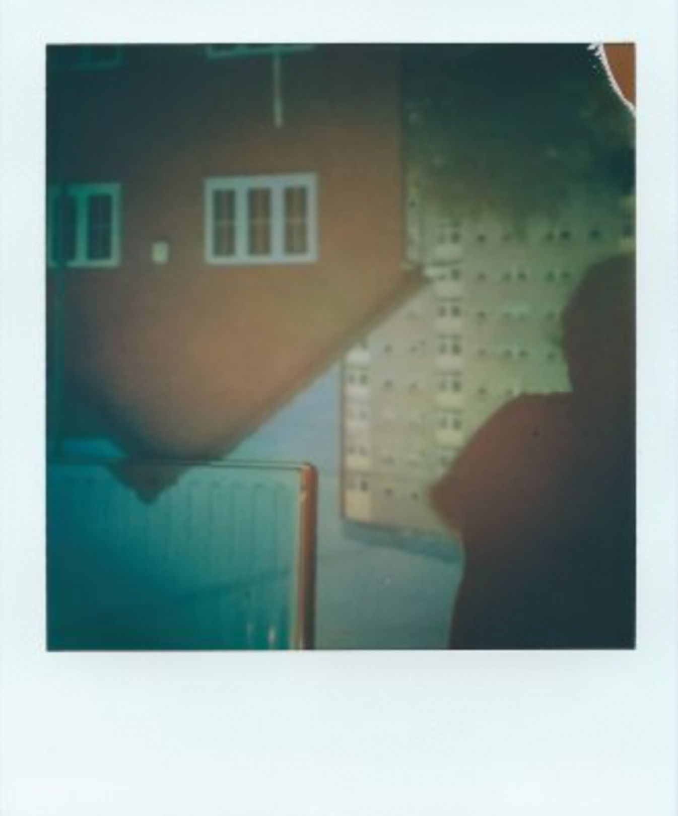 A polaroid image of an upside-down image of a two storey house and high-rise block of flats. This image appears upside-down onto an inside wall, where the top of the roof and a telephone wire cuts onto the room’s radiator. A shadow of a person obscures the right side of the image. The colours are mostly burnt oranges, cream and hints of blue