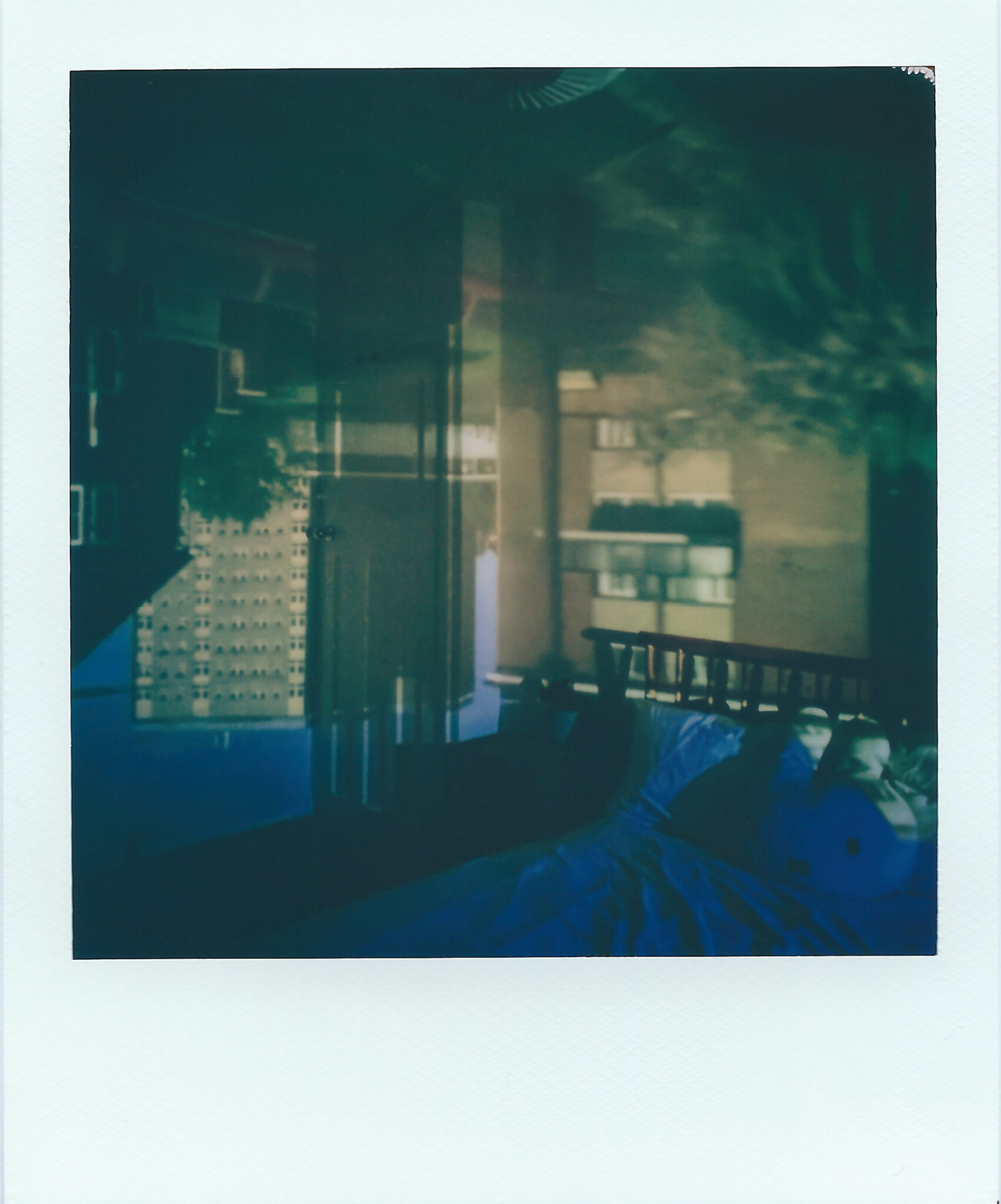 A polaroid image of a busy suburban street that appears upside-down within a bedroom. The flats cover two walls, and the blue sky overlaps the bed. The colours are mostly browns, cream and hints of blue.