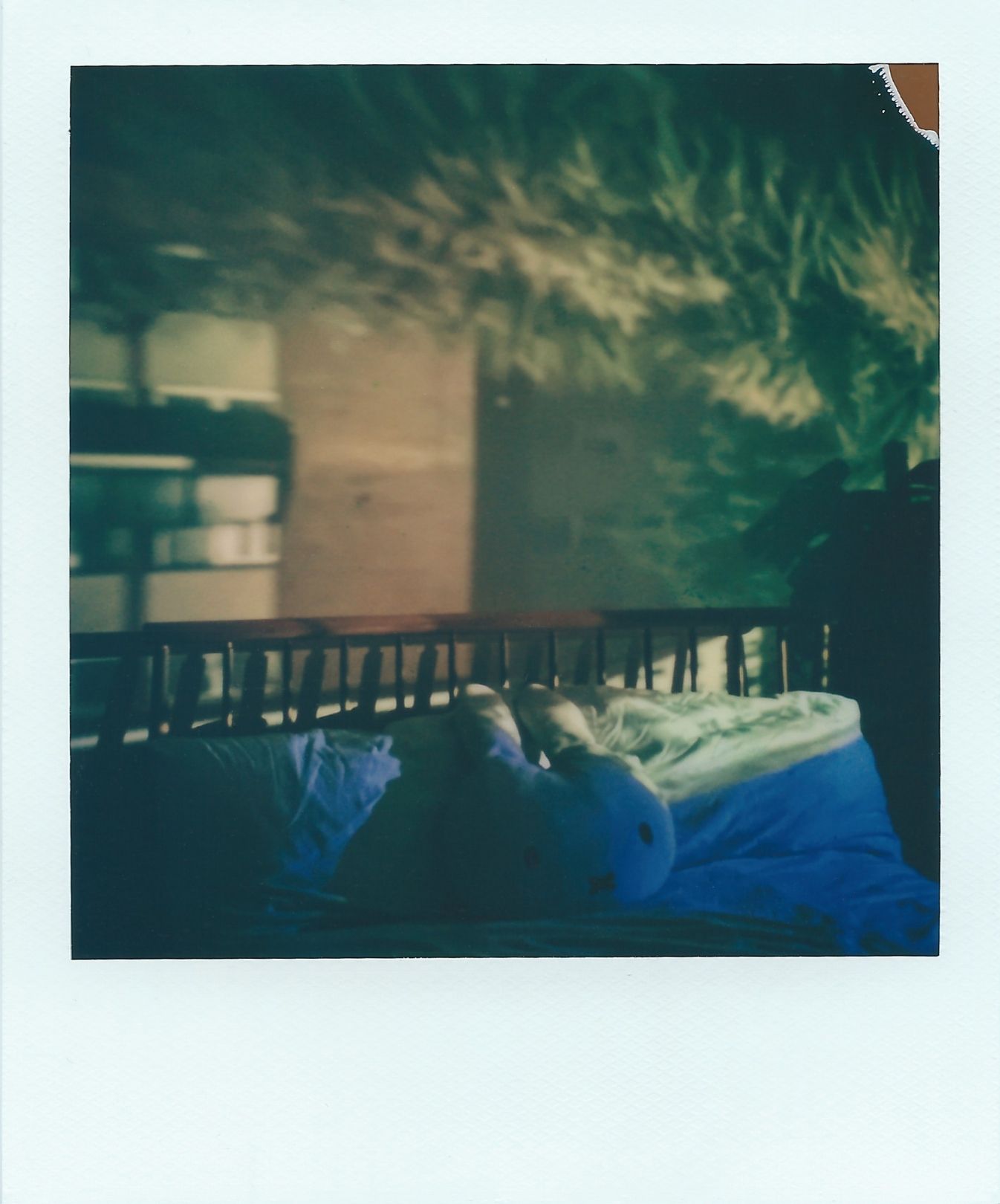 A polaroid image of a flat window and vegetation appears upside-down within a bedroom. The image covers one wall, and the blue sky overlaps the bed. The colours are mostly browns, green and hints of blue and cream.