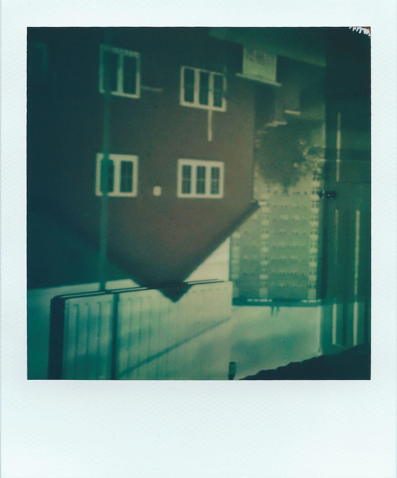 A polaroid image of an upside-down image of a two storey house and high-rise block of flats. This image appears upside-down on an inside wall, where the top of the roof and a lamppost cuts onto the room’s radiator. The flats cut into an interior door. The colours are mostly browns, cream and hints of blue.