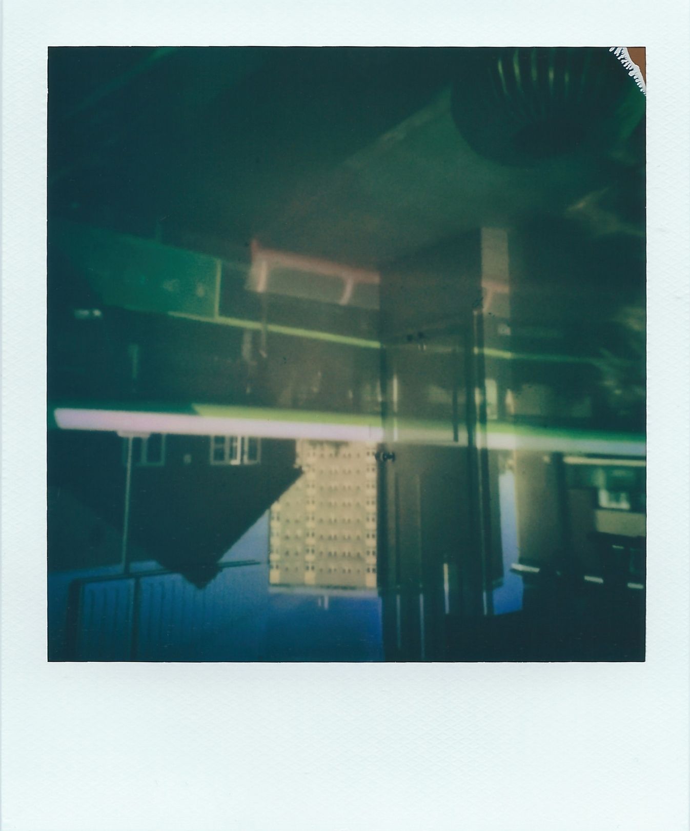 A polaroid image of a busy suburban street that appears upside-down within a bedroom. The flats cover two walls. A bus travels horizontally through the centre of the image, so a streak of green and white cuts across the image with colours that are mostly browns, cream and hints of blue.