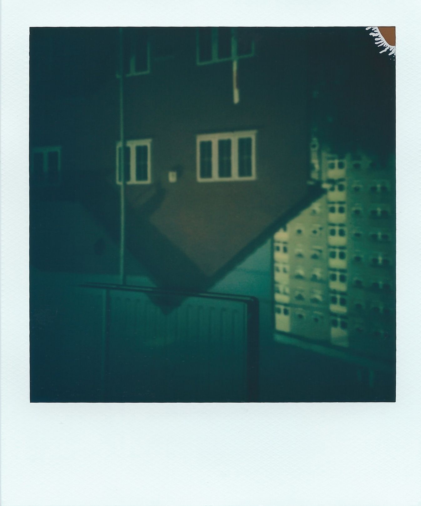A polaroid image of an upside-down image of a two storey house and high-rise block of flats. This image appears upside-down onto an inside wall, where the top of the roof and a lamppost cuts onto the room’s radiator. The colours are mostly browns, cream and hints of blue.