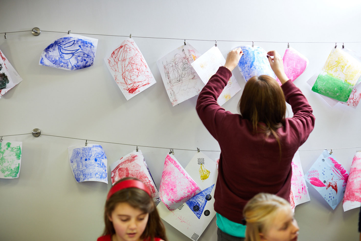 two children in the foreground, a person in the background hands sheets of paper with colour abstract shapes and patterns on two wires