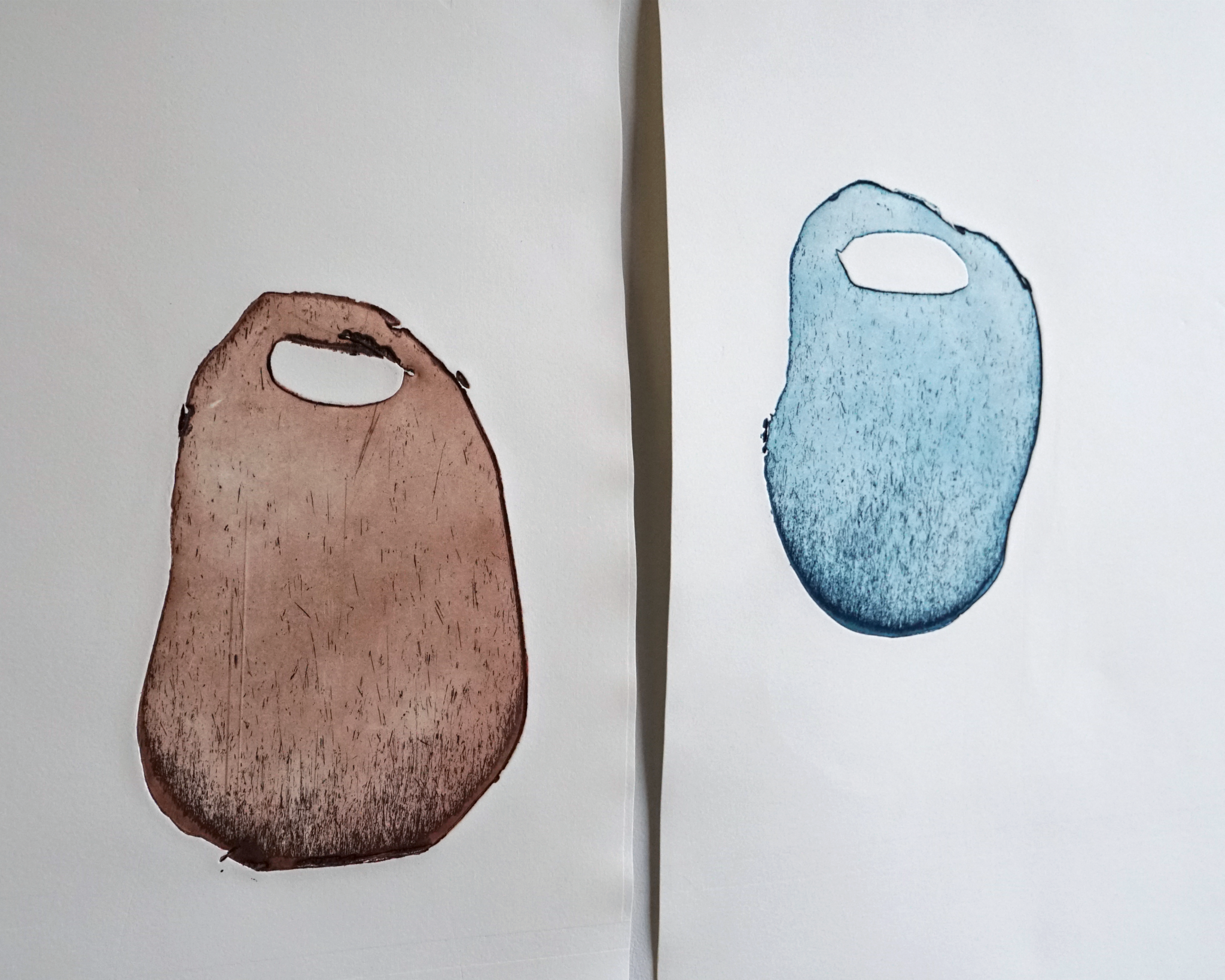 Two prints from metal plates side by side. One is printed in rusty-brown and one is printed in blue. The prints are similar but slightly different in shape. A shadow from the right hand print splits the image in half.