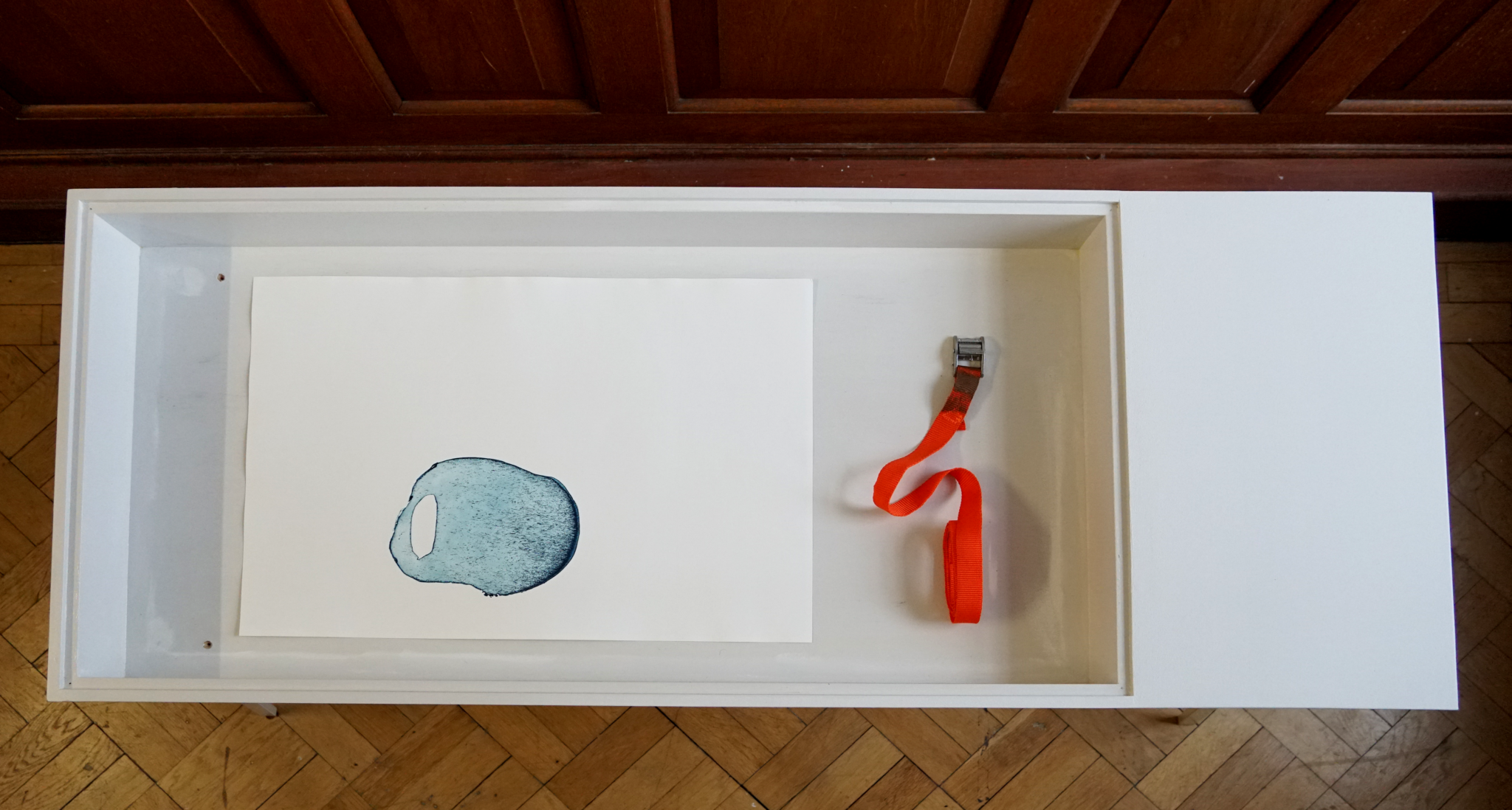 Wide shot looking down on a white display cabinet, which contains a piece of paper with a print from a metal plate in blue and a neon orange ratchet strap.