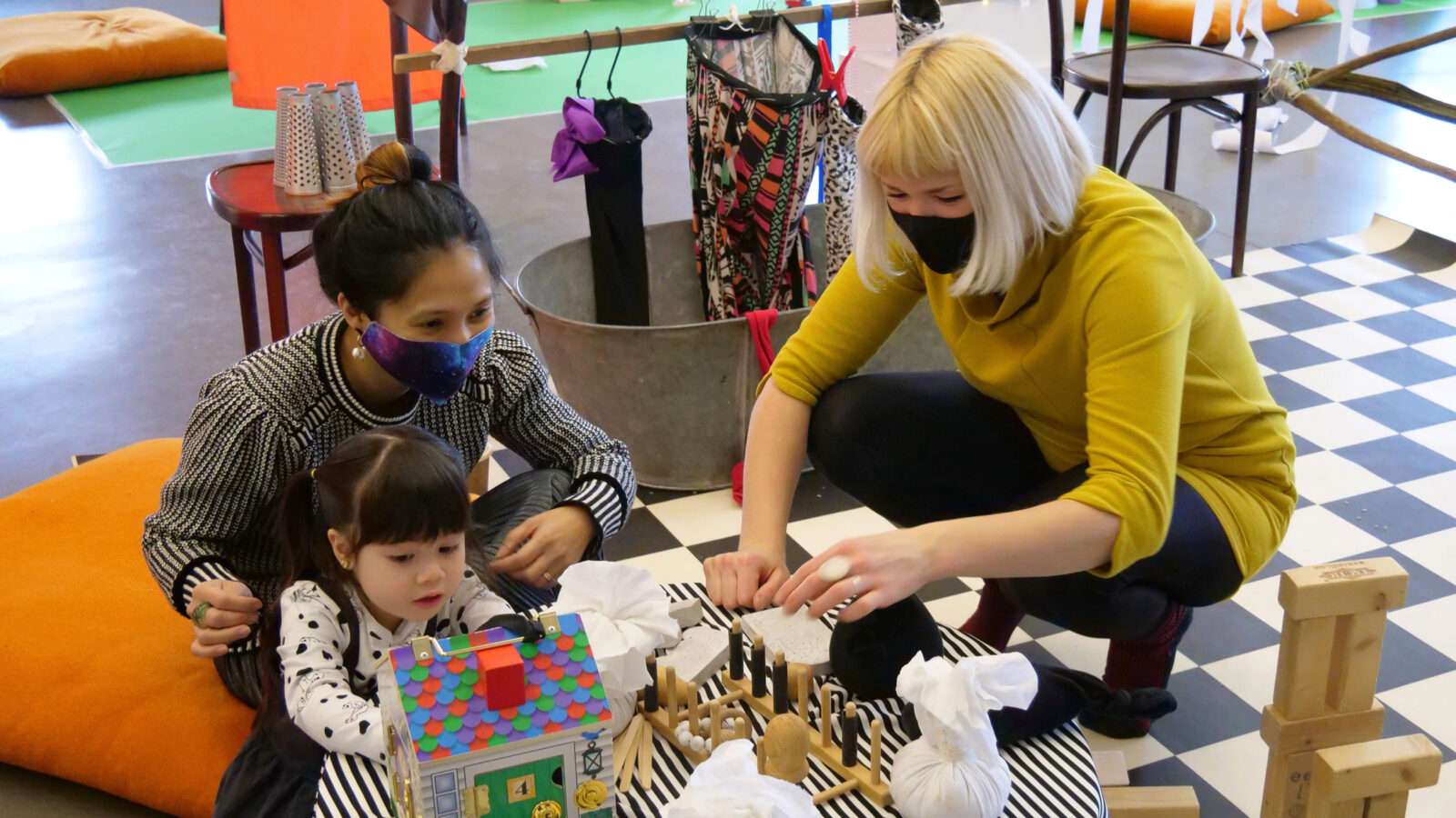 A child and two adults in a workshop with objects including a toy house