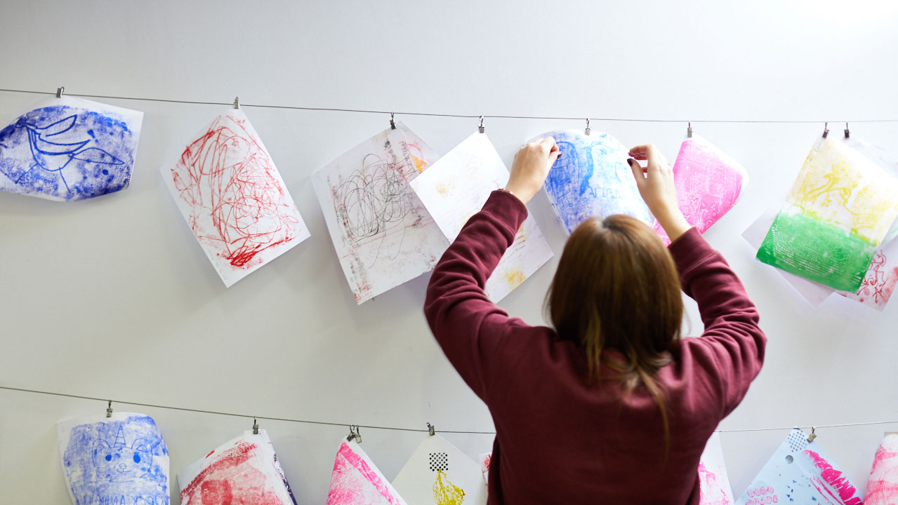 A person in a maroon jacket hanging pieces of paper with brightly coloured abstract shapes on them on a string