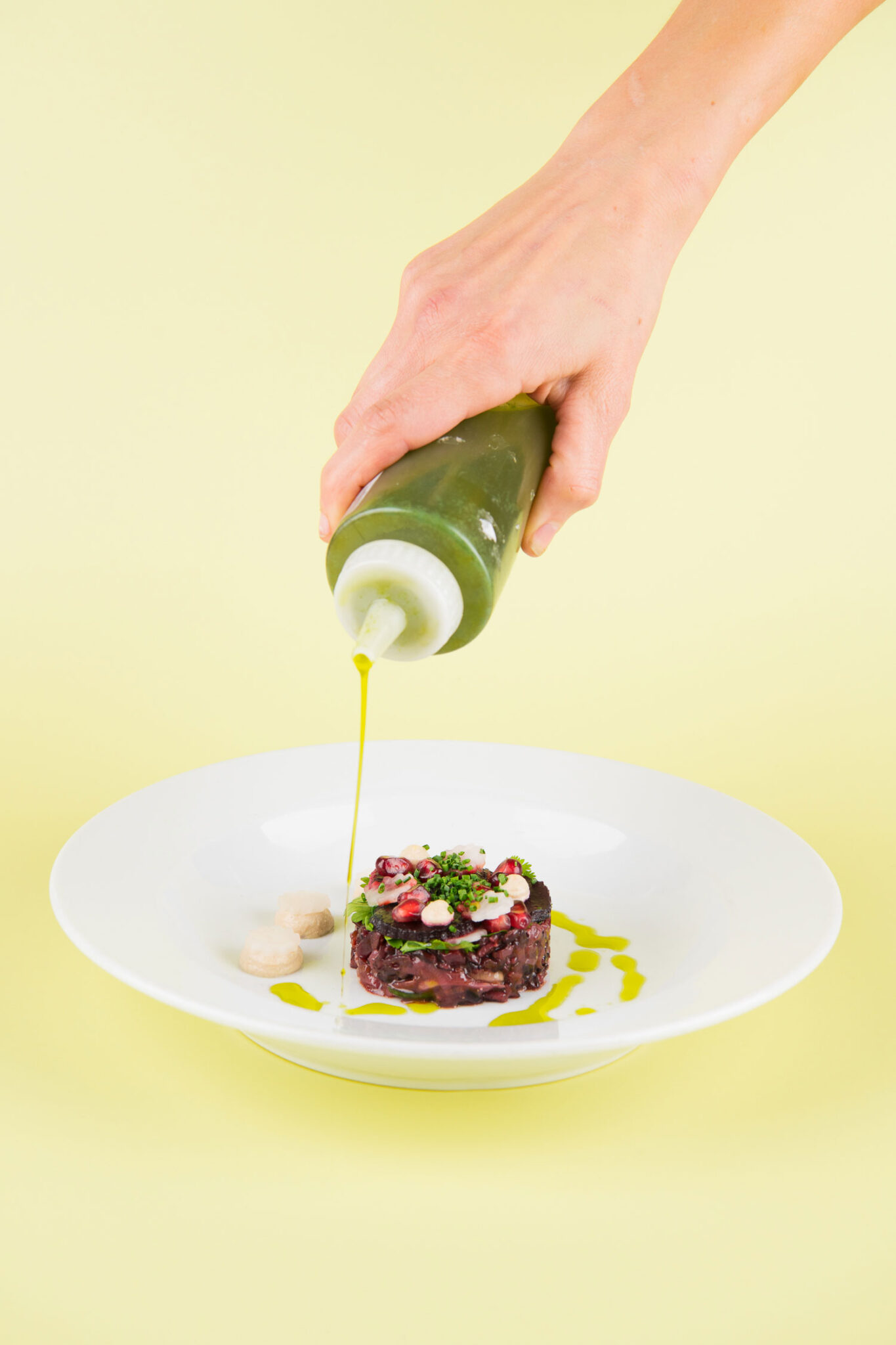 A hand squeezes a bottle of green sauce onto a dish of beetroot and wild rice salad with pomegranate seeds on top