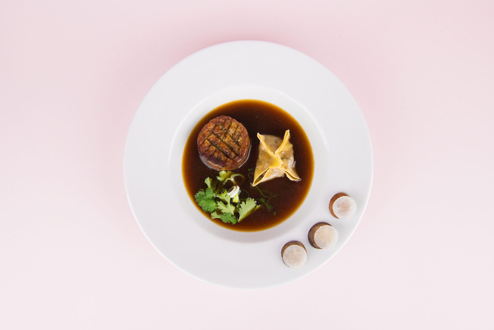 A pale pink background with a white plate, featuring a dumpling and a mushroom in a deep brown broth