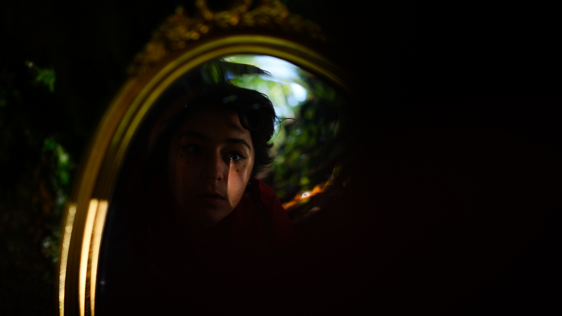 Darkness in most of the image accept for a mirror who reflects a woman’s face half in darkness, with a beam of light on her right cheek