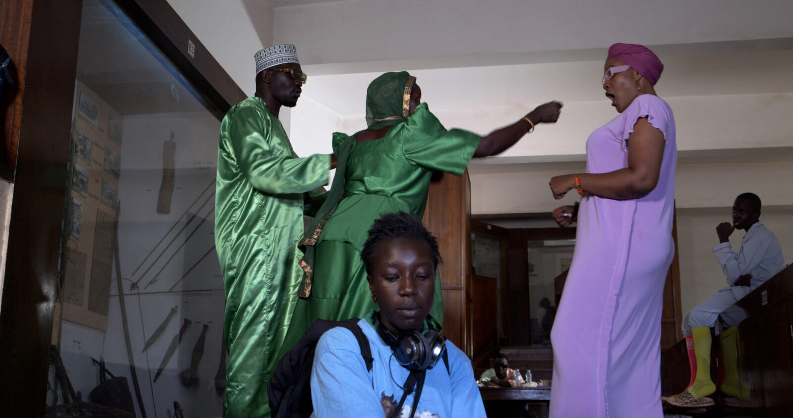 A film still with the artist in the forefront with eyes downcast