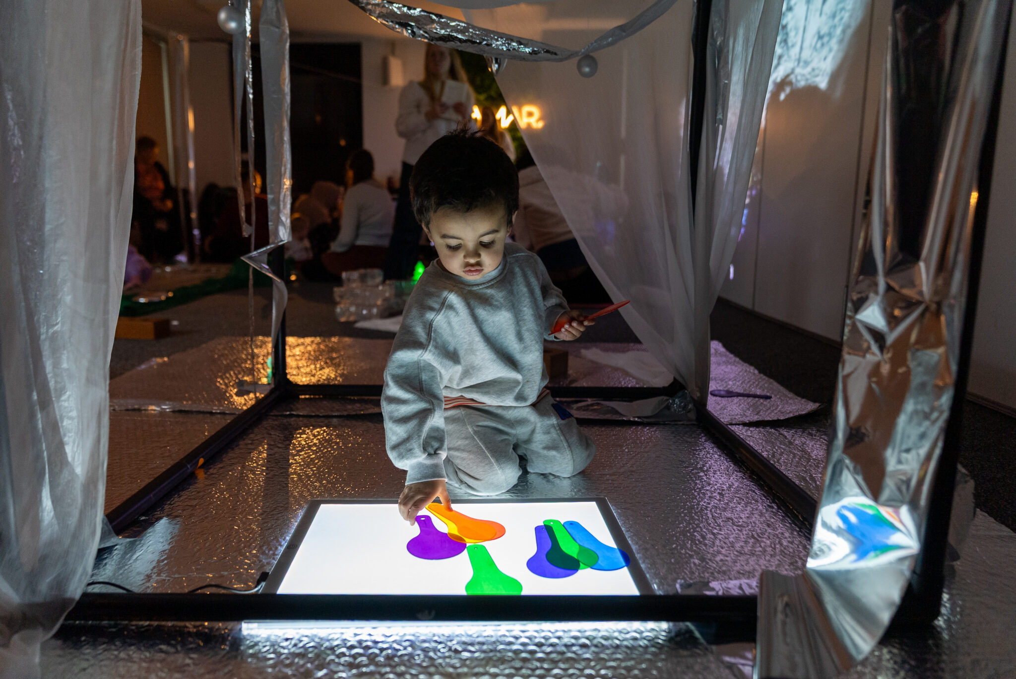 A child in a grey outfit interacts with a brightly lit color panel, touching colorful shapes in a dimly lit, reflective room filled with people.