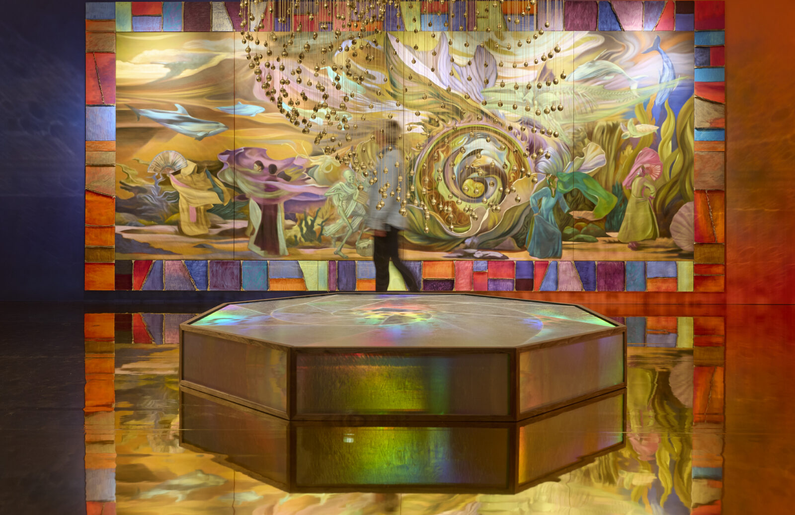 A person walks past a colorful, swirling mural with abstract figures and marine life, reflected on a gold shiny floor. In front, delicate, conch like shells dangle from the ceiling above a raised hexagonal plinth.The scene is vibrant and dreamlike.