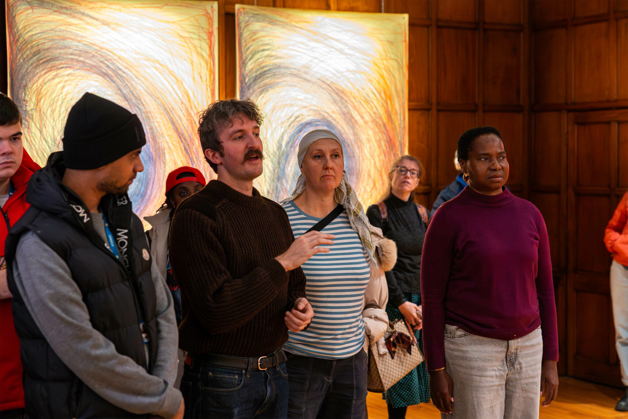 A diverse group of people stand attentively in an art gallery, with vibrant, swirling abstract paintings in the background, creating a thoughtful atmosphere.