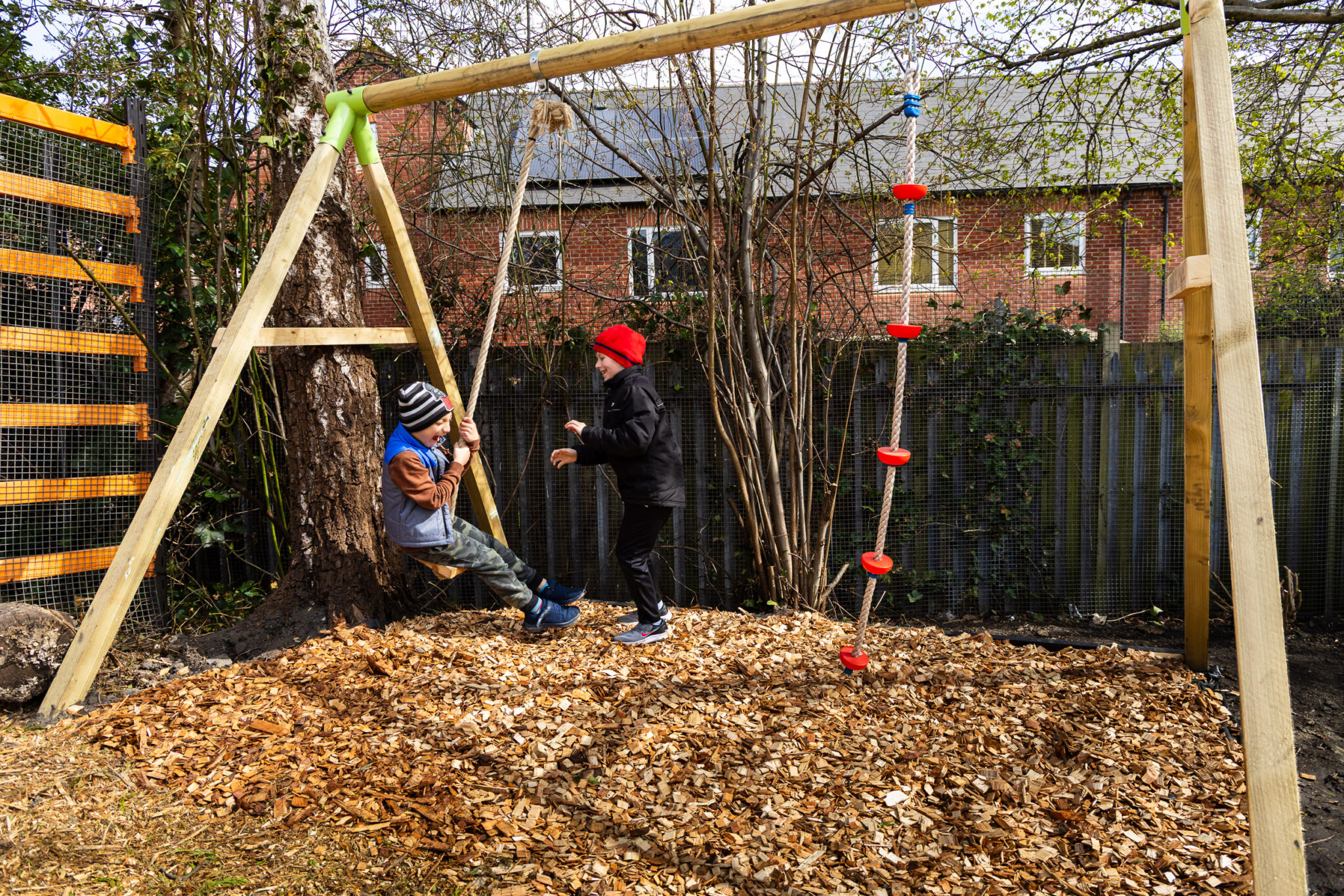 Two children play on a wooden swing set with bark mulch groundcover. One swings while the other stands nearby, both smiling. Trees and a brick house are in the background.