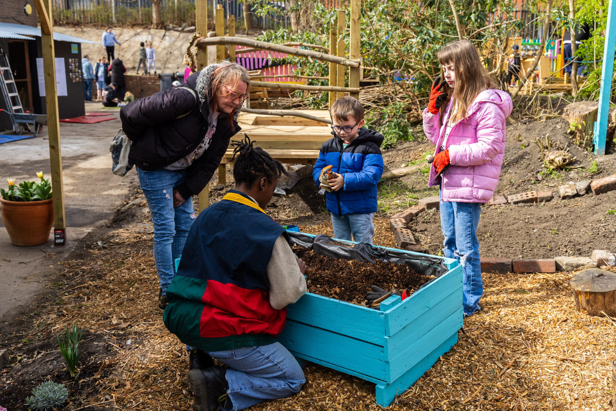 A person crouches by a blue planter, engaging with two children and an adult. The children are bundled in colorful jackets. It's a joyful garden scene.