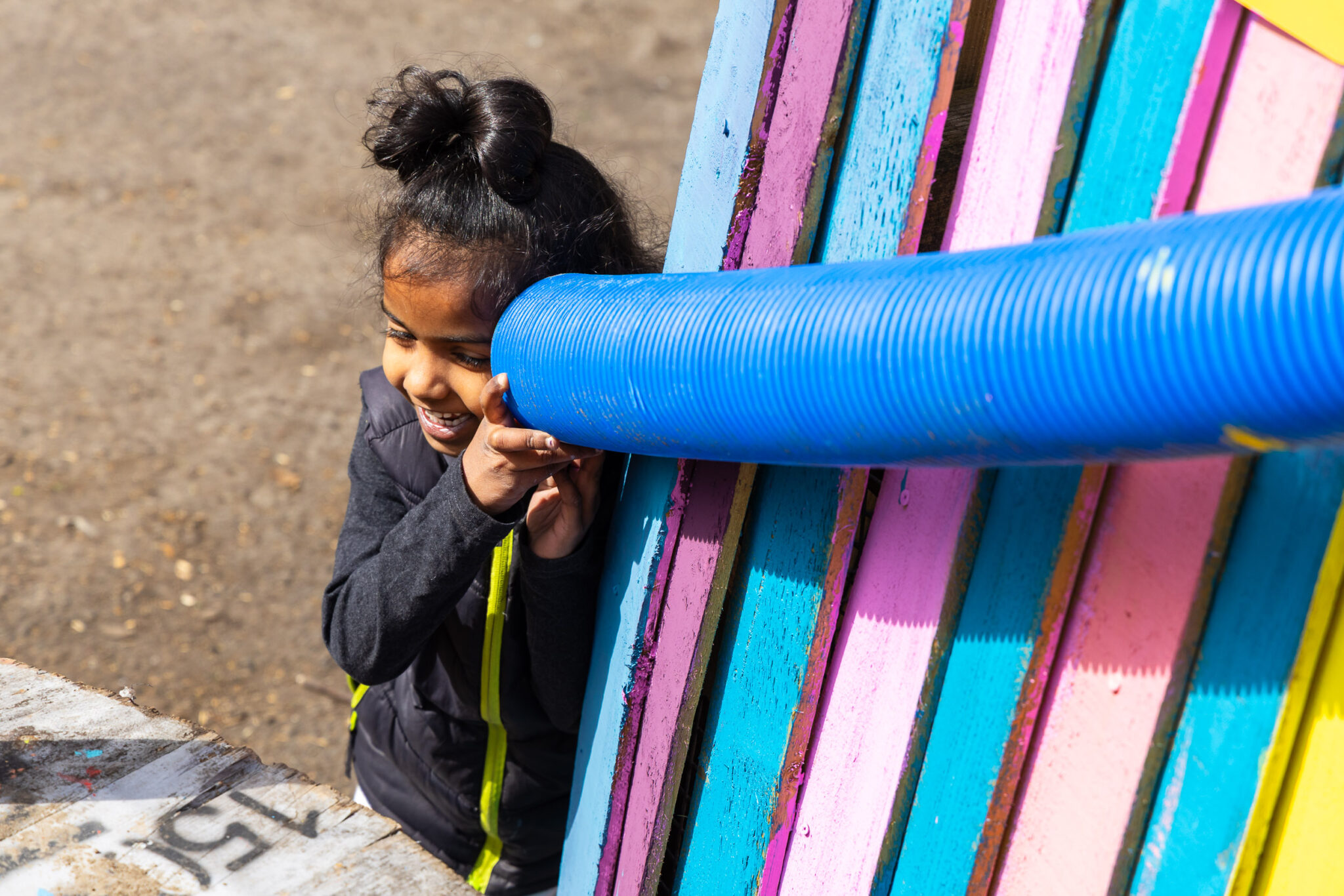 A child with hair in a bun smiles and listens through a blue tube on a colorful fence in bright sunlight. The scene conveys joy and playfulness.