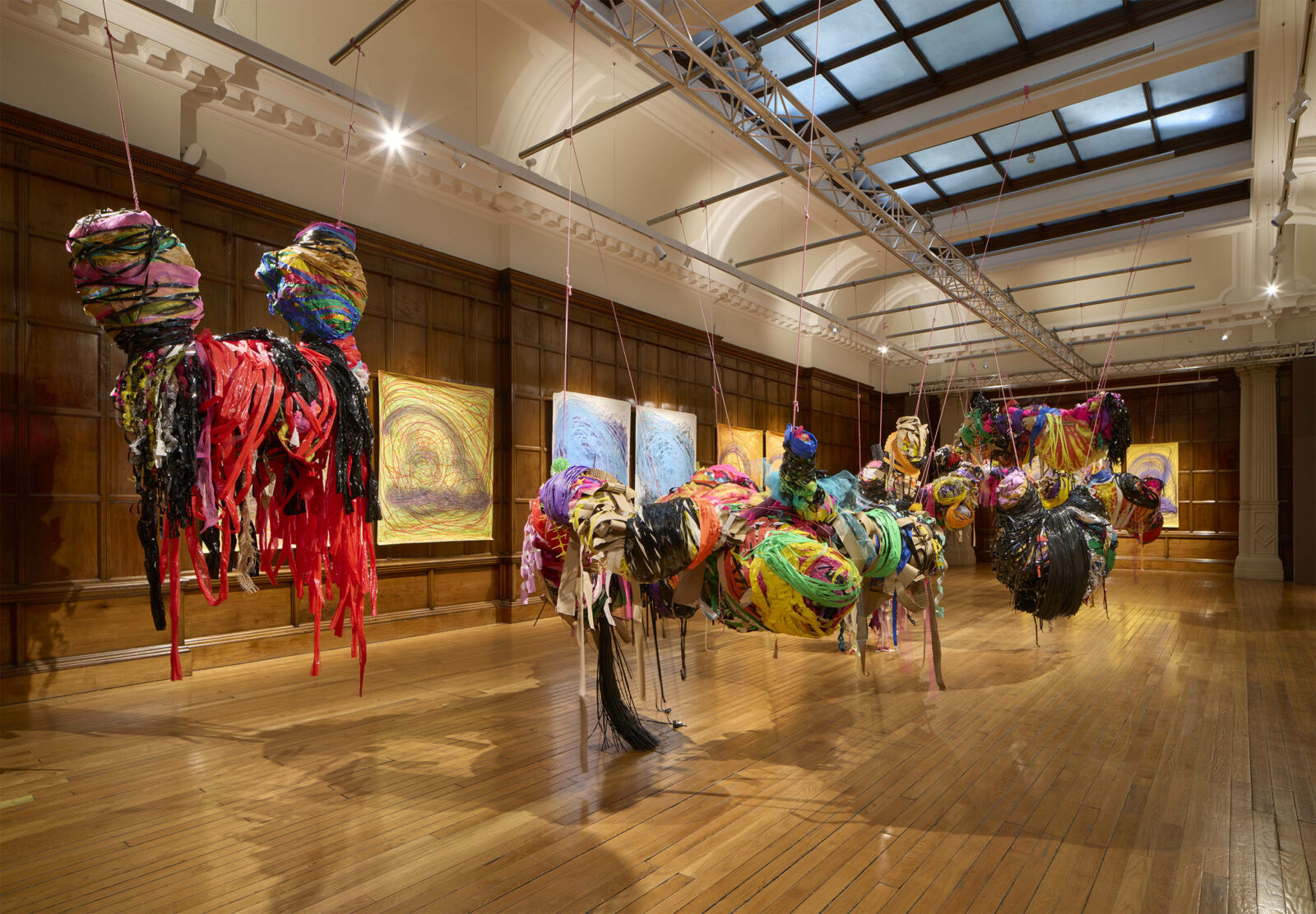 Installation view of Nnena Kalu’s presentation at Turner Prize 2025 in a wood-panelled gallery. Large, suspended sculptural forms made from brightly coloured, layered and wrapped materials hang from the ceiling, stretching across the room, with vibrant abstract paintings displayed on the walls behind them.