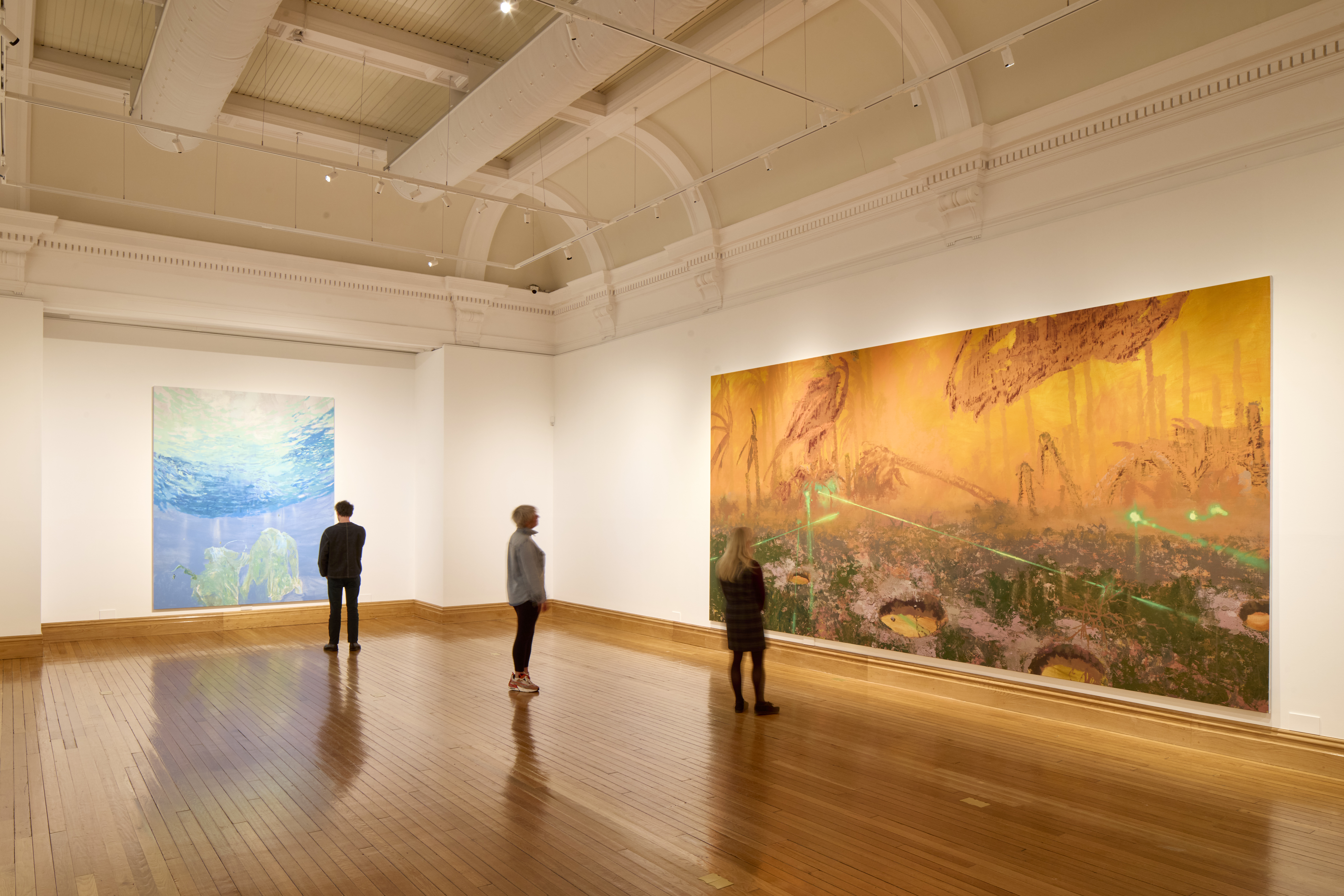 A large, high-ceilinged art gallery with polished wooden floors and white walls. Two oversized paintings hang on adjacent walls: on the left, a blue-toned underwater scene with jellyfish; on the right, a wide orange and yellow abstract landscape with green lines and textured details. Three visitors stand spaced apart, quietly viewing the artworks under bright overhead lights.