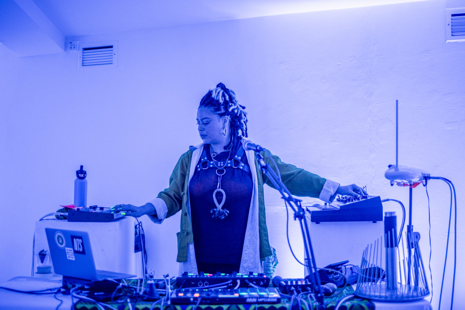 Person with braids in a dimly lit room, surrounded by electronic music equipment. They wear a necklace and look focused, creating an atmospheric vibe.