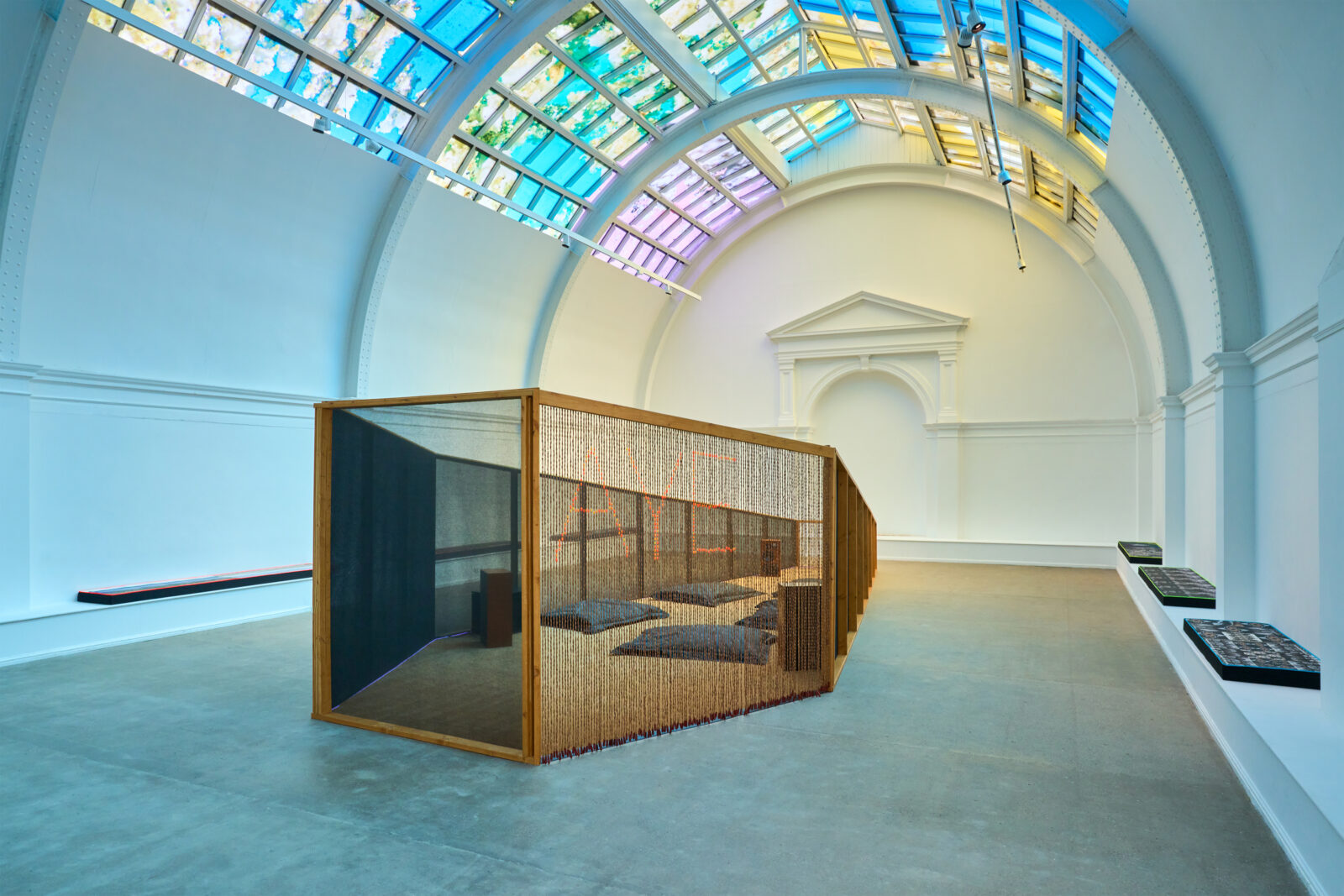A large white room with a colourful lit ceiling above a wooden coffin shaped oversized sculpture.