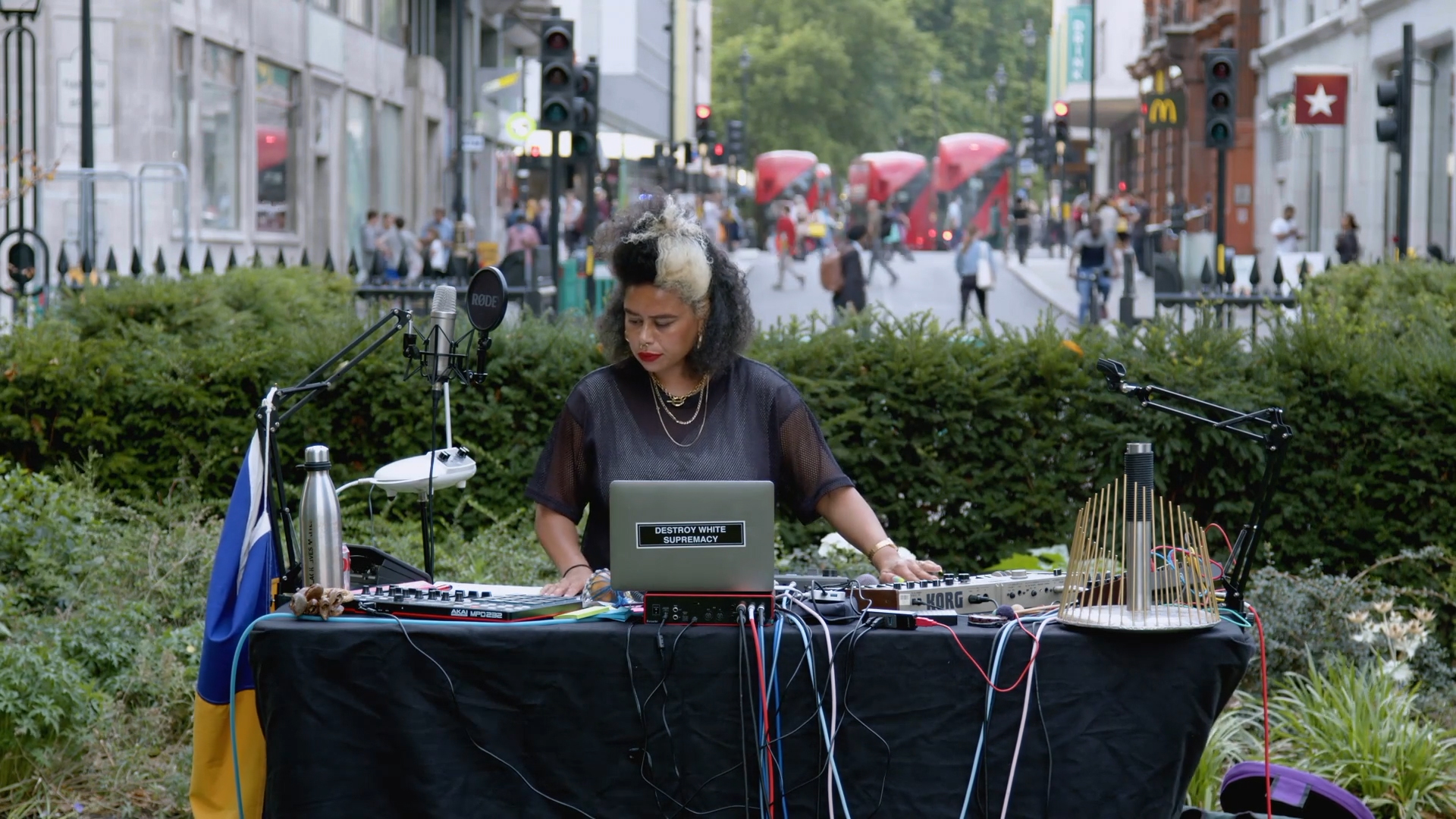 A DJ performs outdoors on a city street, surrounded by music equipment and cables, with a flag draped on the table. Red buses and pedestrians fill the background, creating an urban atmosphere.