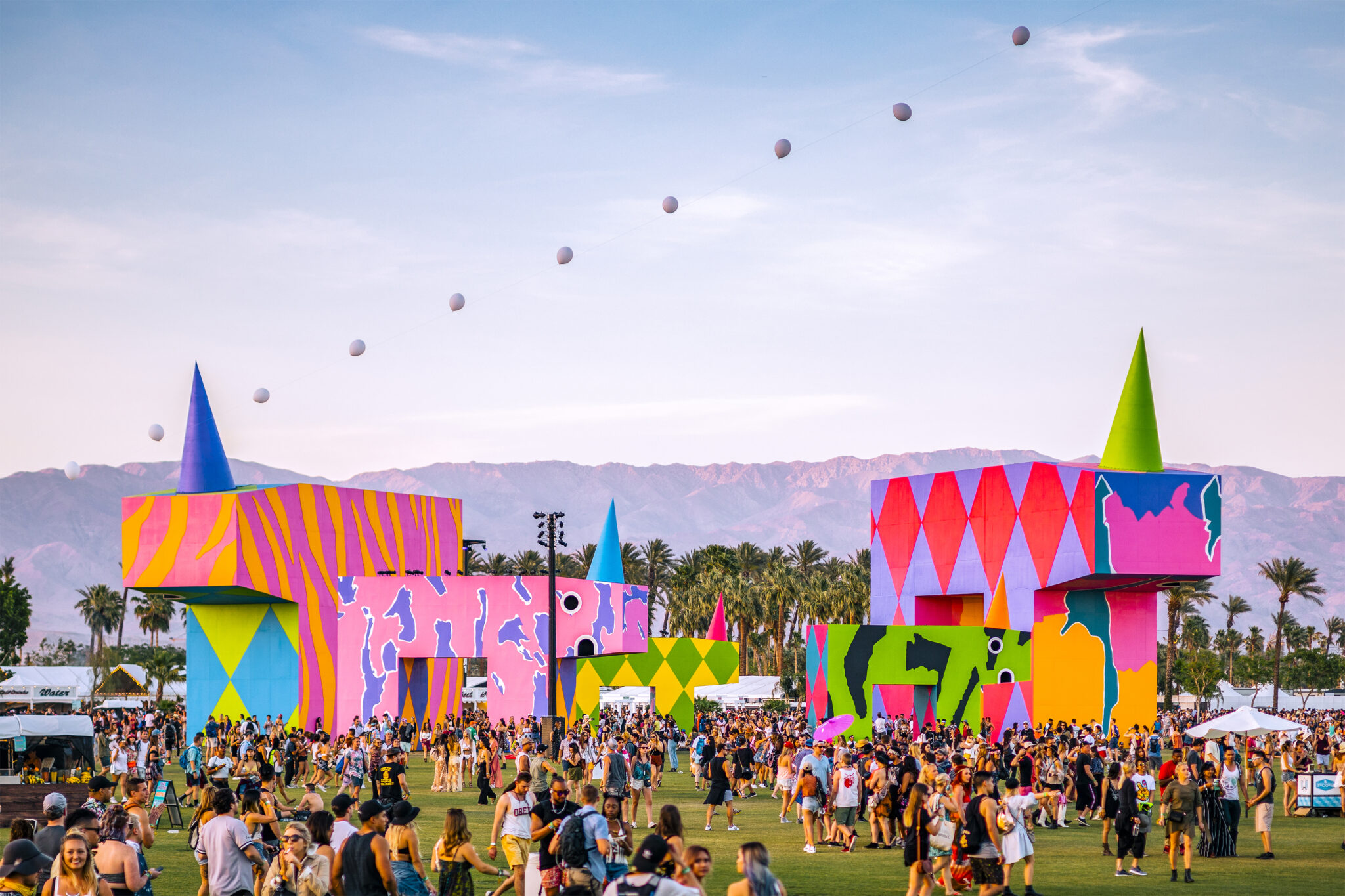 Festival scene with vibrant, geometric art structures and crowds on grassy field. Mountains and clear sky in the background evoke a lively, festive atmosphere.
