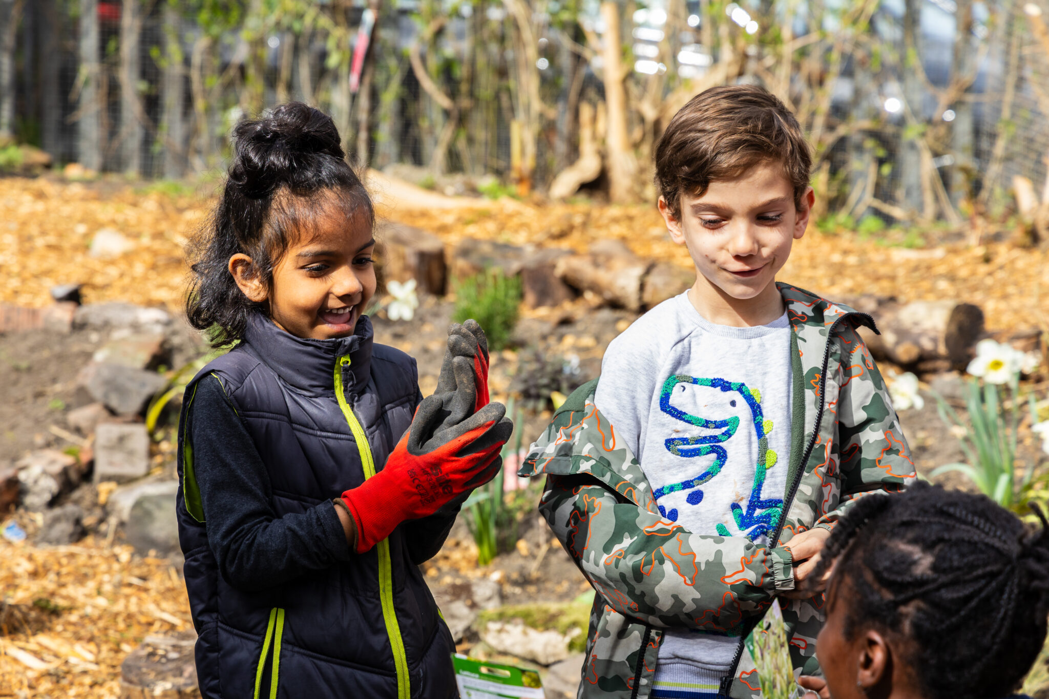 Two children smiling during outdoor gardening activity, wearing gloves and casual jackets, standing in a school garden with soil beds, rocks, and spring flowers in background; kids participating in environmental education, hands-on learning, teamwork, and nature exploration in sunny park setting