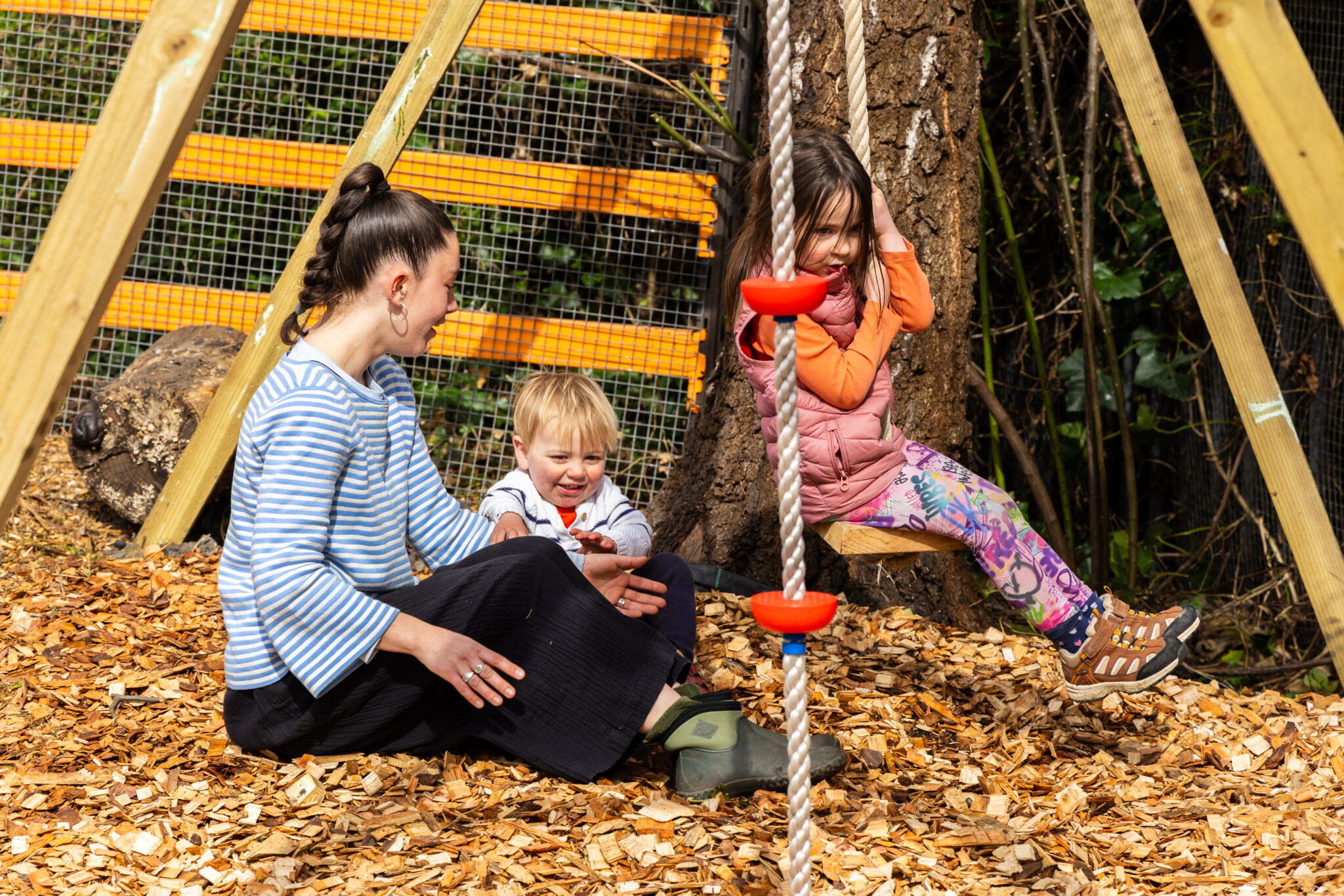 Adult caregiver sitting on wood chips beside two young children playing on an outdoor playground rope swing attached to a tree, natural wooden climbing frame with mesh fencing and logs in background, kids enjoying active play in forest school setting, family time outdoors, early childhood development, safe playground environment