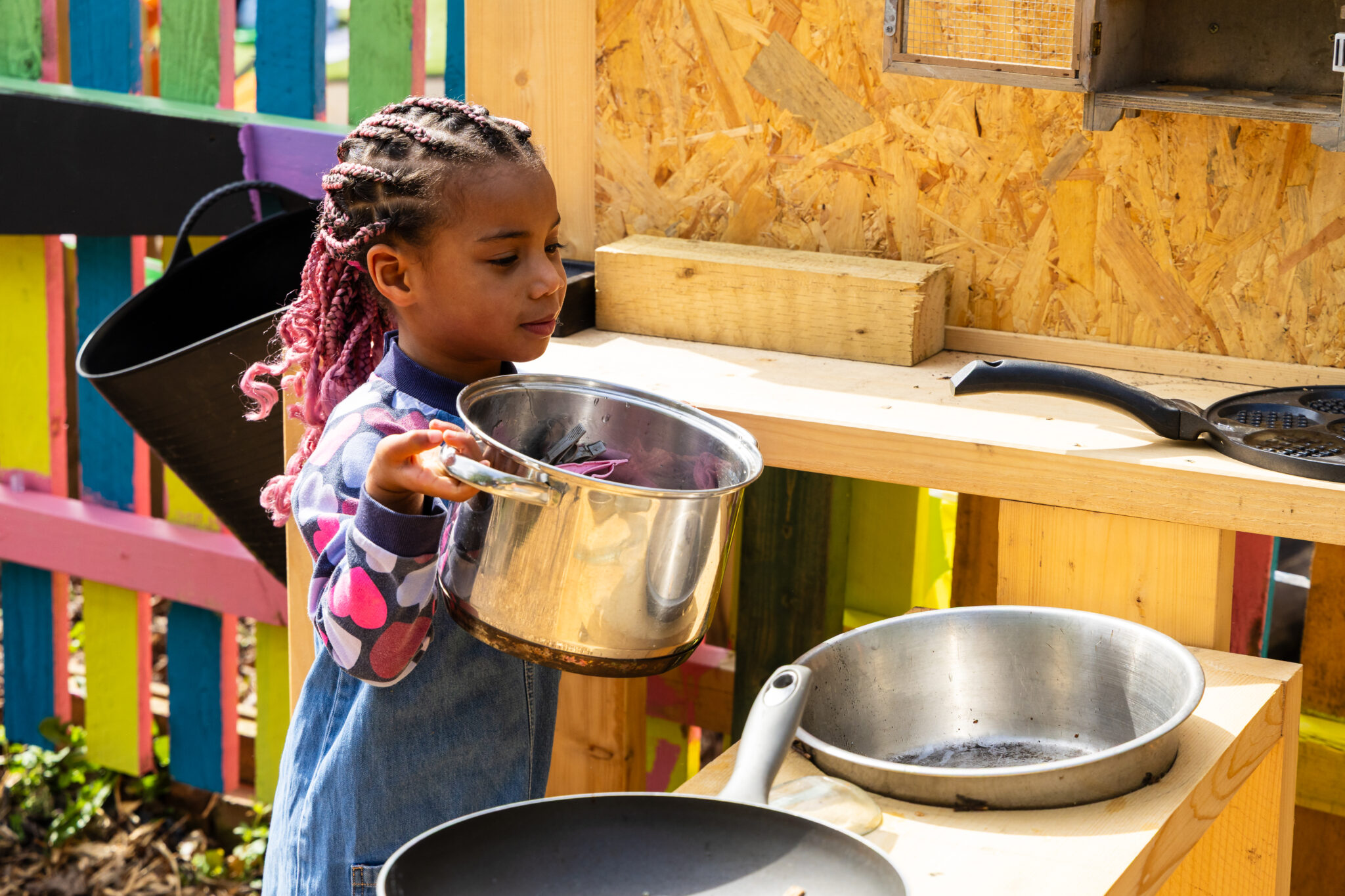 Young child with pink braided hair playing in an outdoor wooden mud kitchen, holding a stainless steel cooking pot over a toy stovetop with metal pans, colorful fence background, pretend cooking activity, early childhood education, creative outdoor play, natural light, backyard playground scene