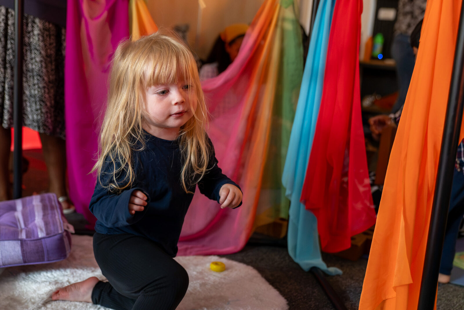 A young child explores an immersive play space surrounded by brightly coloured hanging fabrics.