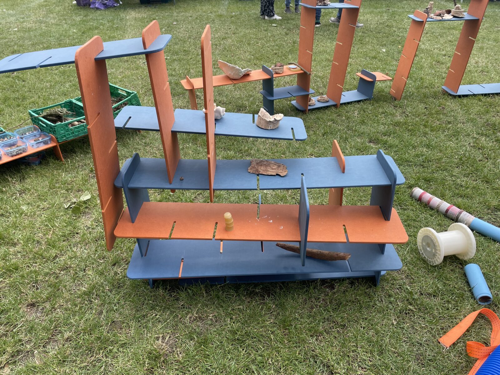 Colorful play structures with orange and blue panels are arranged on grass. Nature items like bark and stones are placed on them. A playful, creative scene.