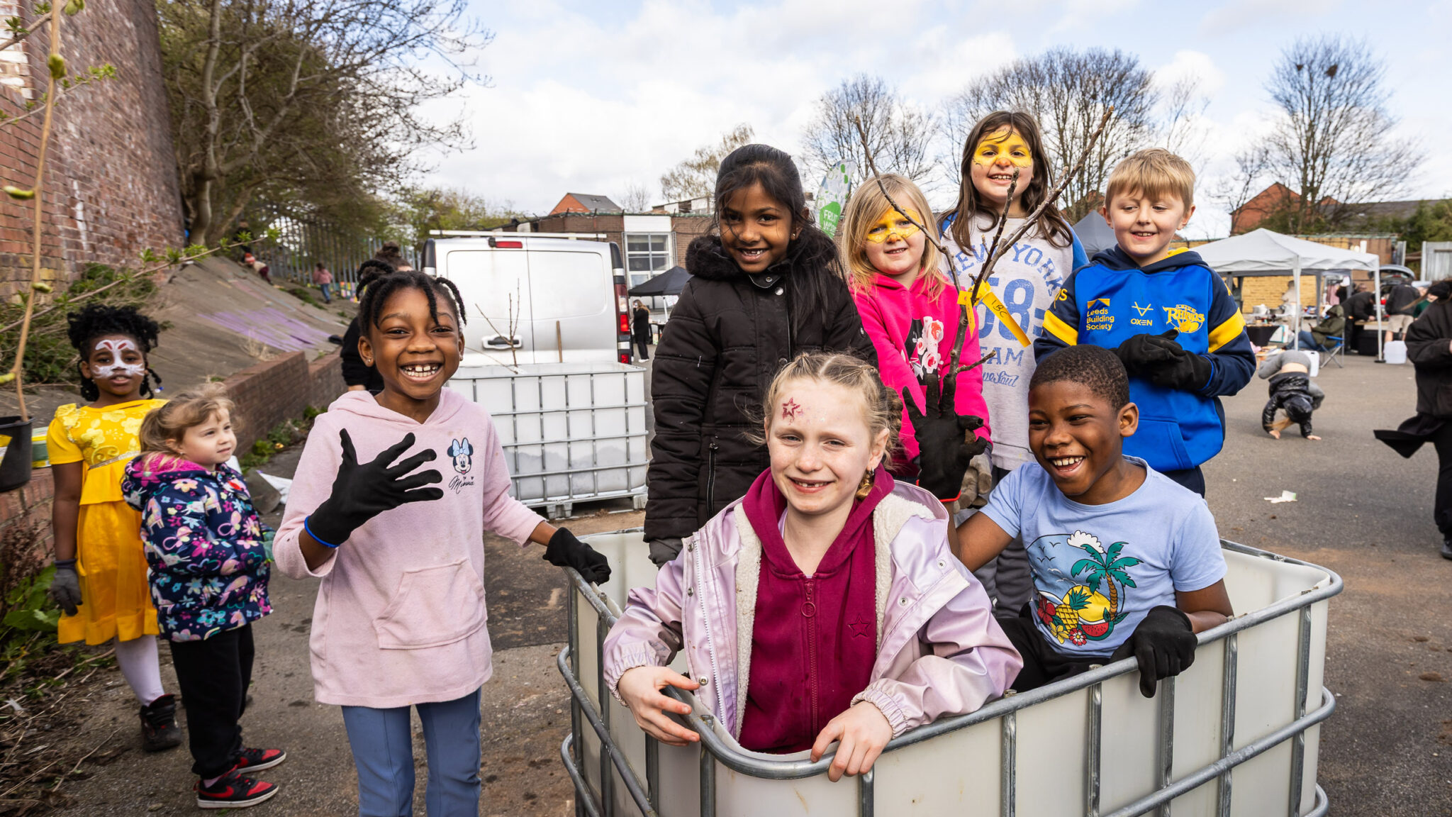 A group of cheerful children in colorful outfits and face paint smile at the camera outdoors, conveying joy and camaraderie on a clear day.