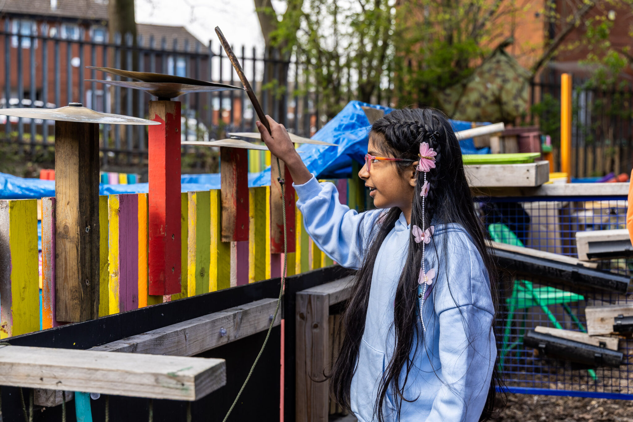 A girl with long hair and a flower clip plays outdoor musical chimes with a stick in a colorful, handmade setting, smiling joyfully.
