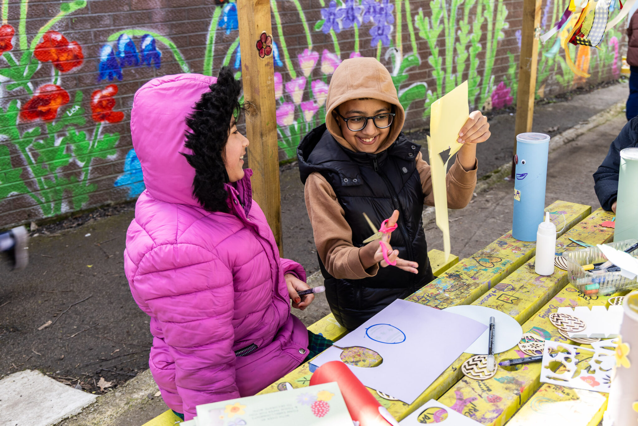 Two children in winter clothing laugh and craft at a colorful table outdoors, with a vibrant floral mural behind them. The scene is joyful and creative.