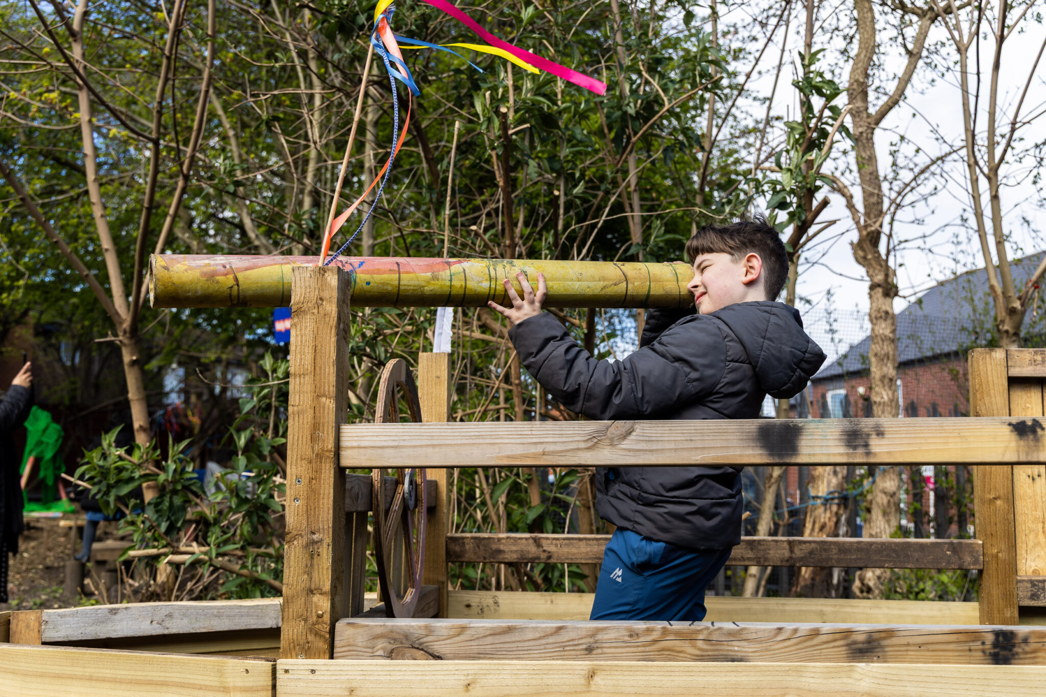 A boy in a black jacket looks through a large bamboo telescope adorned with colorful ribbons, set in a wooden play structure in a leafy garden.