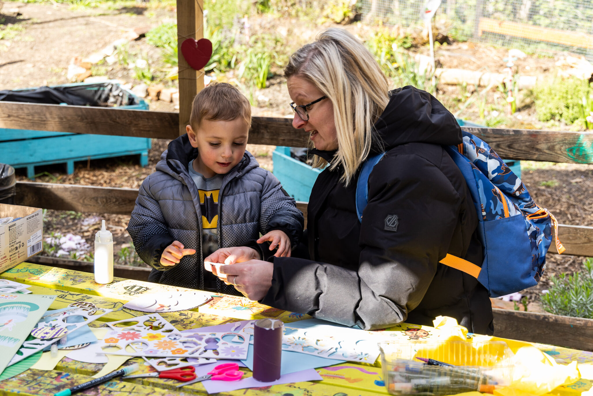 A mother and son smile while crafting at an outdoor table covered with colorful paper and art supplies. The setting is bright and cheerful.