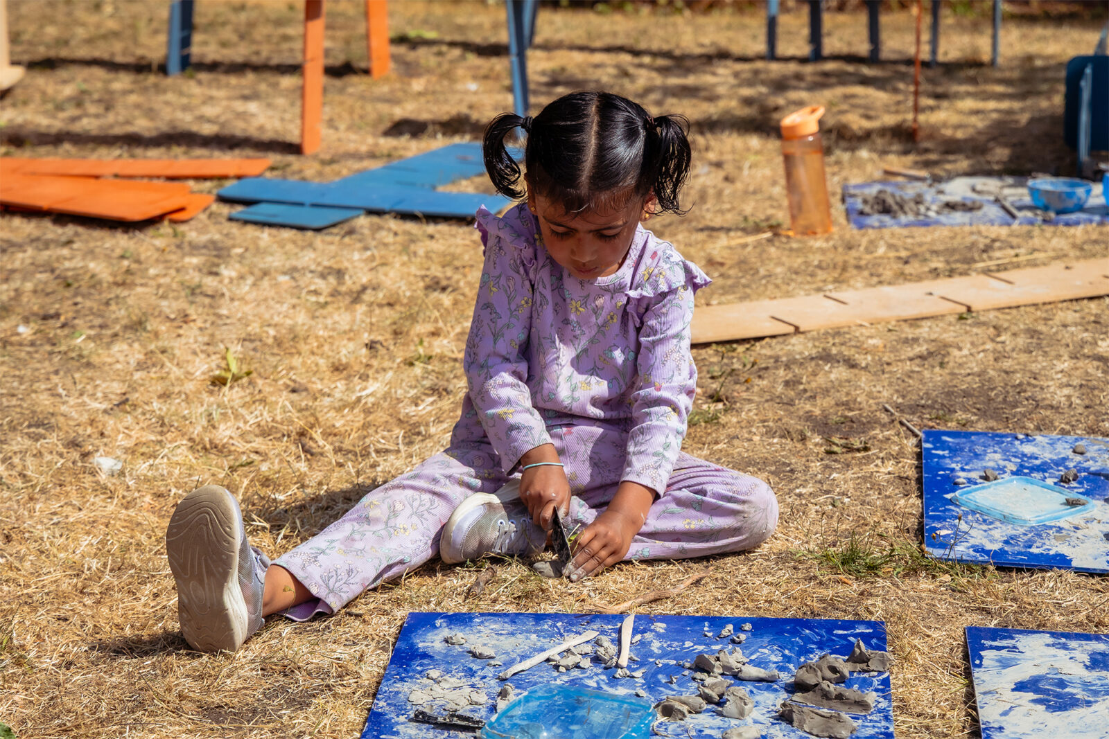 A young child in a purple floral outfit sits on dry grass, shaping pieces of grey clay on a blue board during an outdoor creative activity.