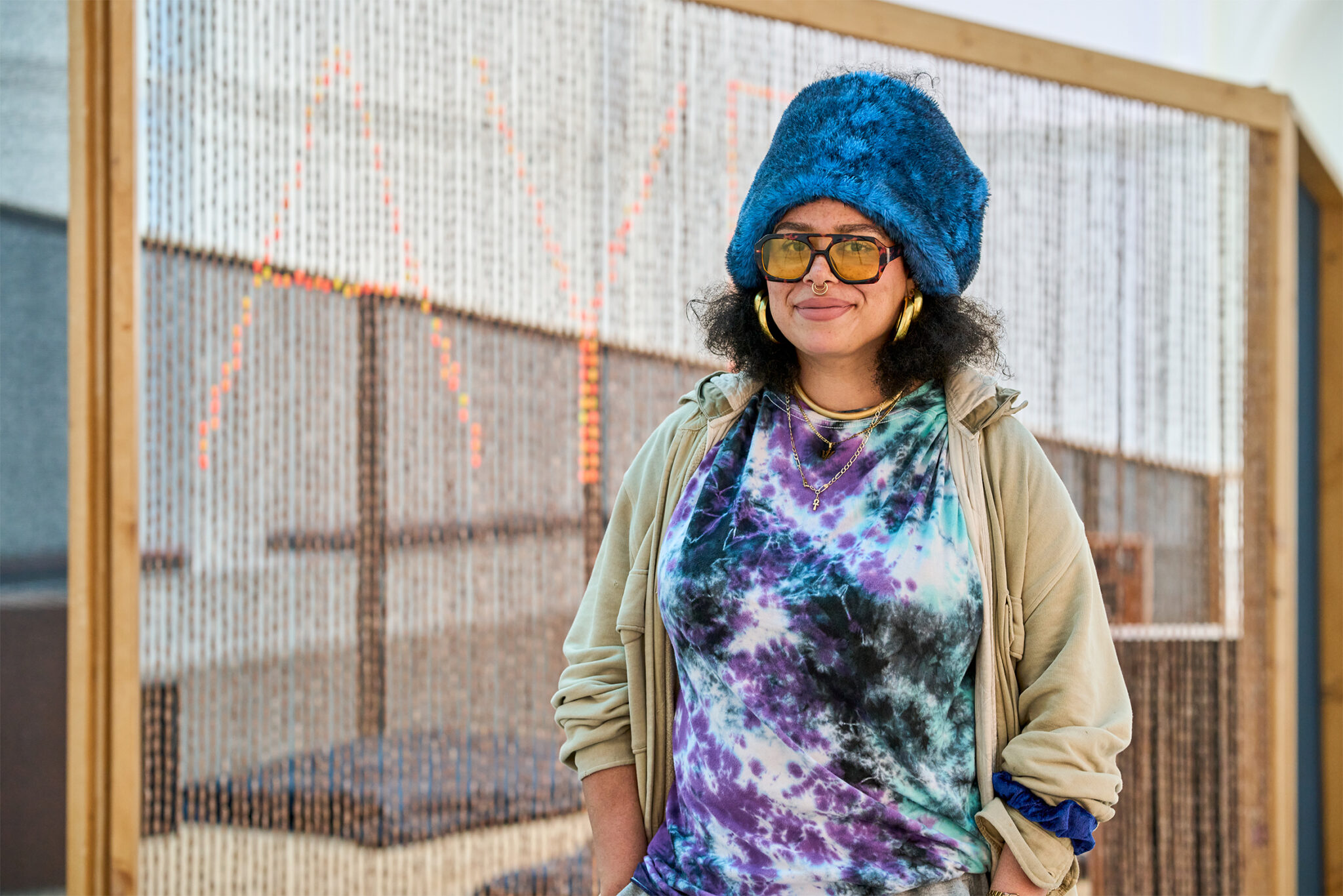 A person stands smiling in front of a large woven installation framed in wood. They wear a bright blue fuzzy hat, oversized tinted glasses, large gold hoop earrings, a nose ring, layered necklaces, a colorful purple, blue, and black tie-dye shirt, and a light beige jacket. Their hands are tucked into their pockets. Behind them, the woven artwork partially reveals glowing orange letters, creating a textured backdrop in a bright gallery space.