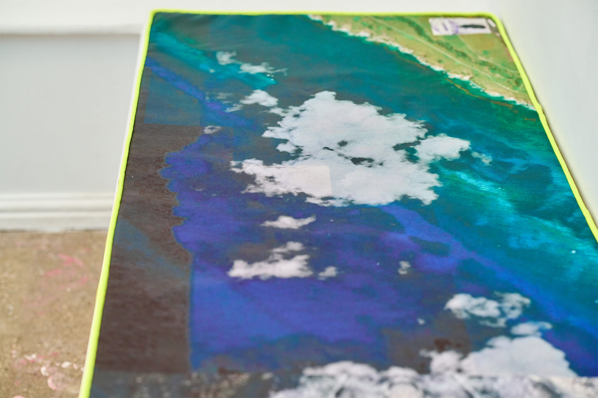 A large rectangular textile or printed fabric lies flat on the gallery floor, depicting an aerial view of a coastline with vivid turquoise and deep blue water, scattered white clouds, and a narrow strip of green land along one edge. The fabric is bordered with a bright fluorescent yellow trim and photographed from a low angle, emphasizing its length within the white-walled gallery space.