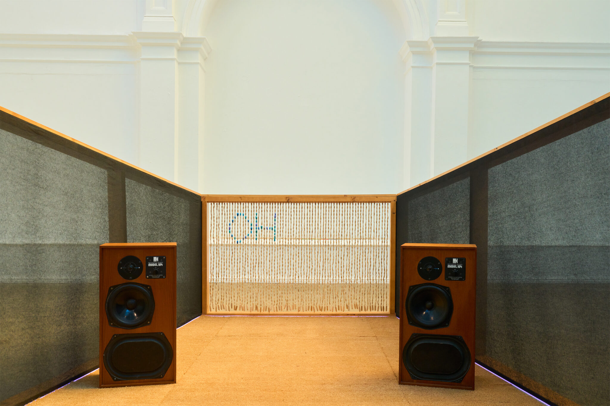 A symmetrical gallery installation featuring two large vintage-style wooden speakers facing down a narrow corridor lined with tall dark acoustic panels. At the far end, a woven screen partially reveals the blue letters 