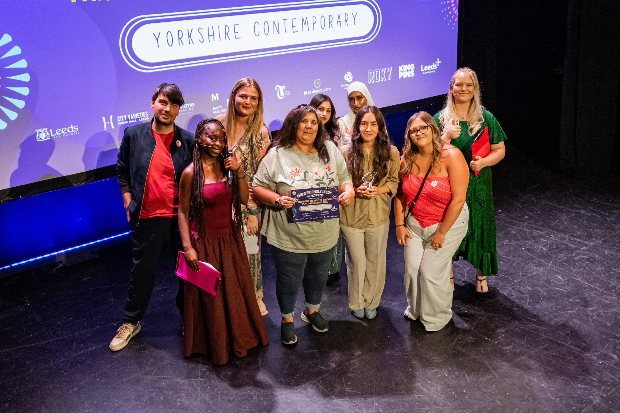 A group of nine people stand on stage celebrating after winning the Inspiring Creativity Through Play, Arts & Culture award at the Child Friendly Leeds Awards 2026. Several people hold the award trophy and certificate while smiling for the camera. Behind them, a large screen displays the Yorkshire Contemporary logo alongside the Child Friendly Leeds Awards branding and sponsor logos. The group includes staff, volunteers and young people involved in the award-winning Rowland Road Play Patch project.